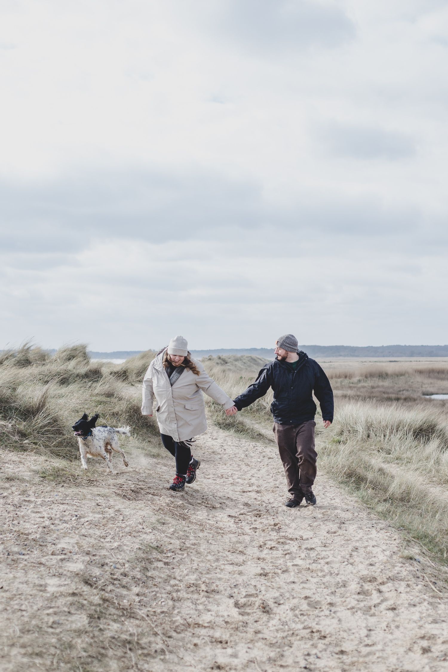 Two people walking with dog on coastal path through sand dunes.