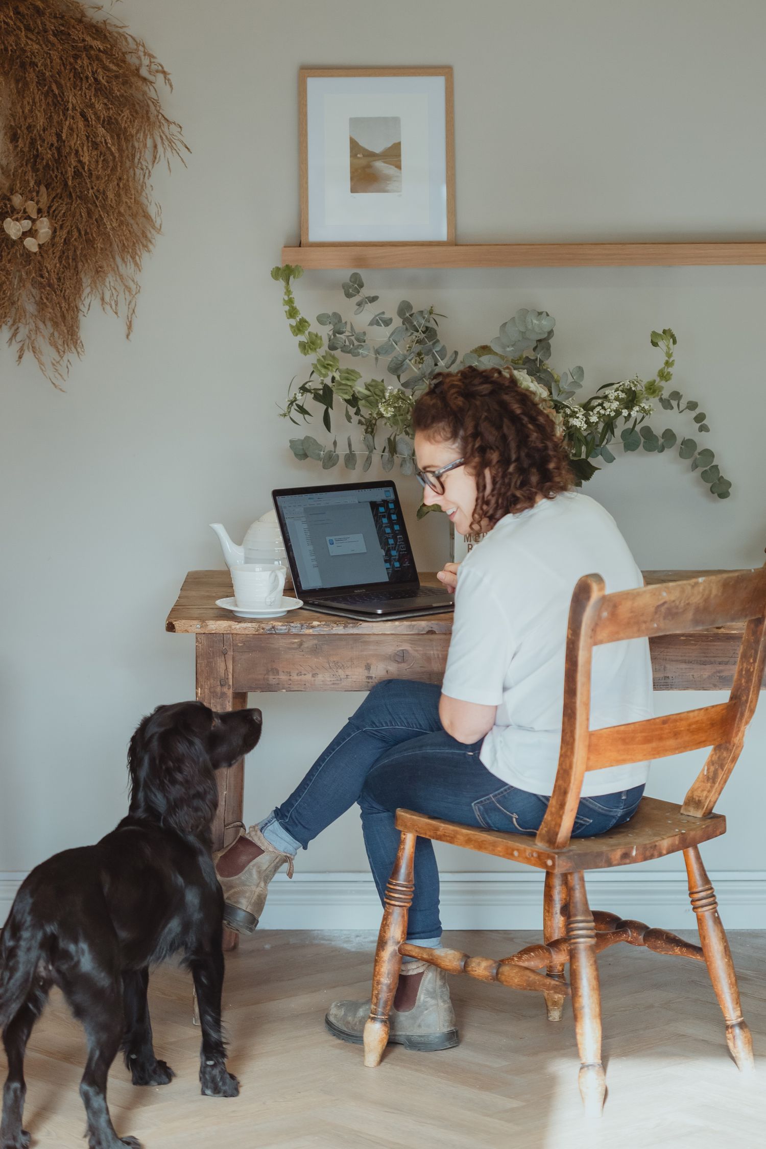 Person working on laptop at wooden desk with black dog sitting beside them.