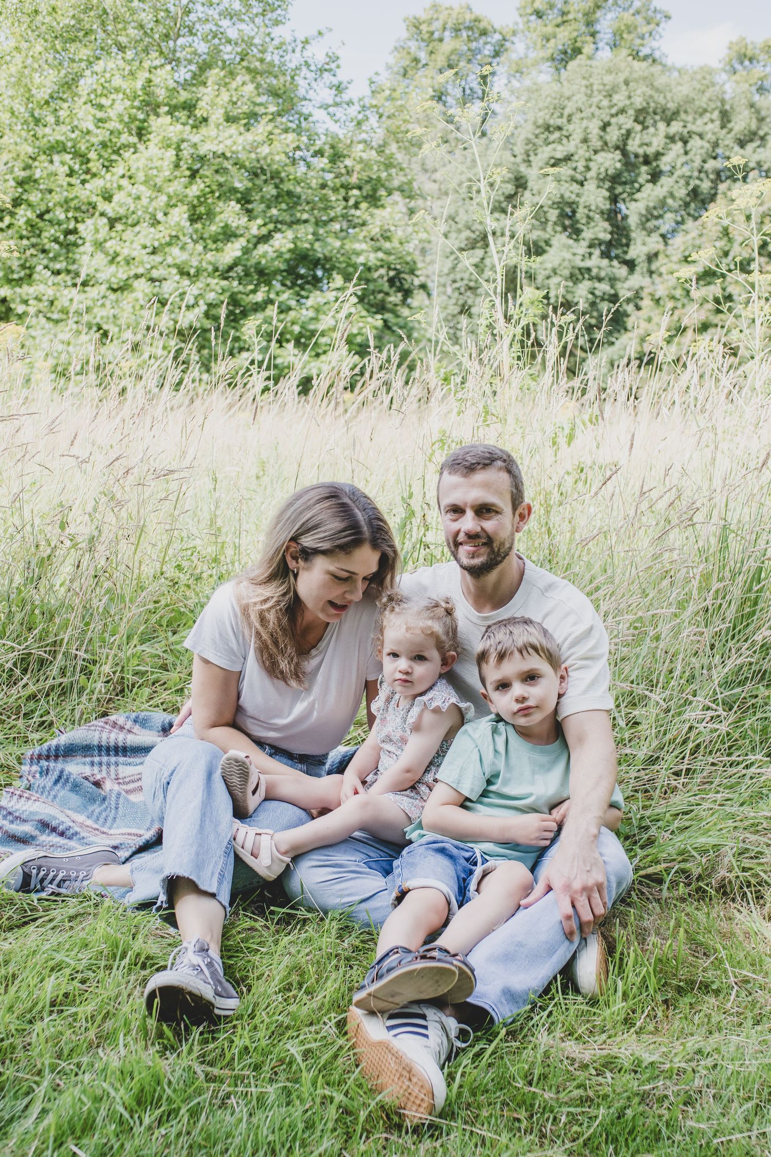 A family relaxes together on a picnic blanket in a sunny meadow with tall grass and trees.