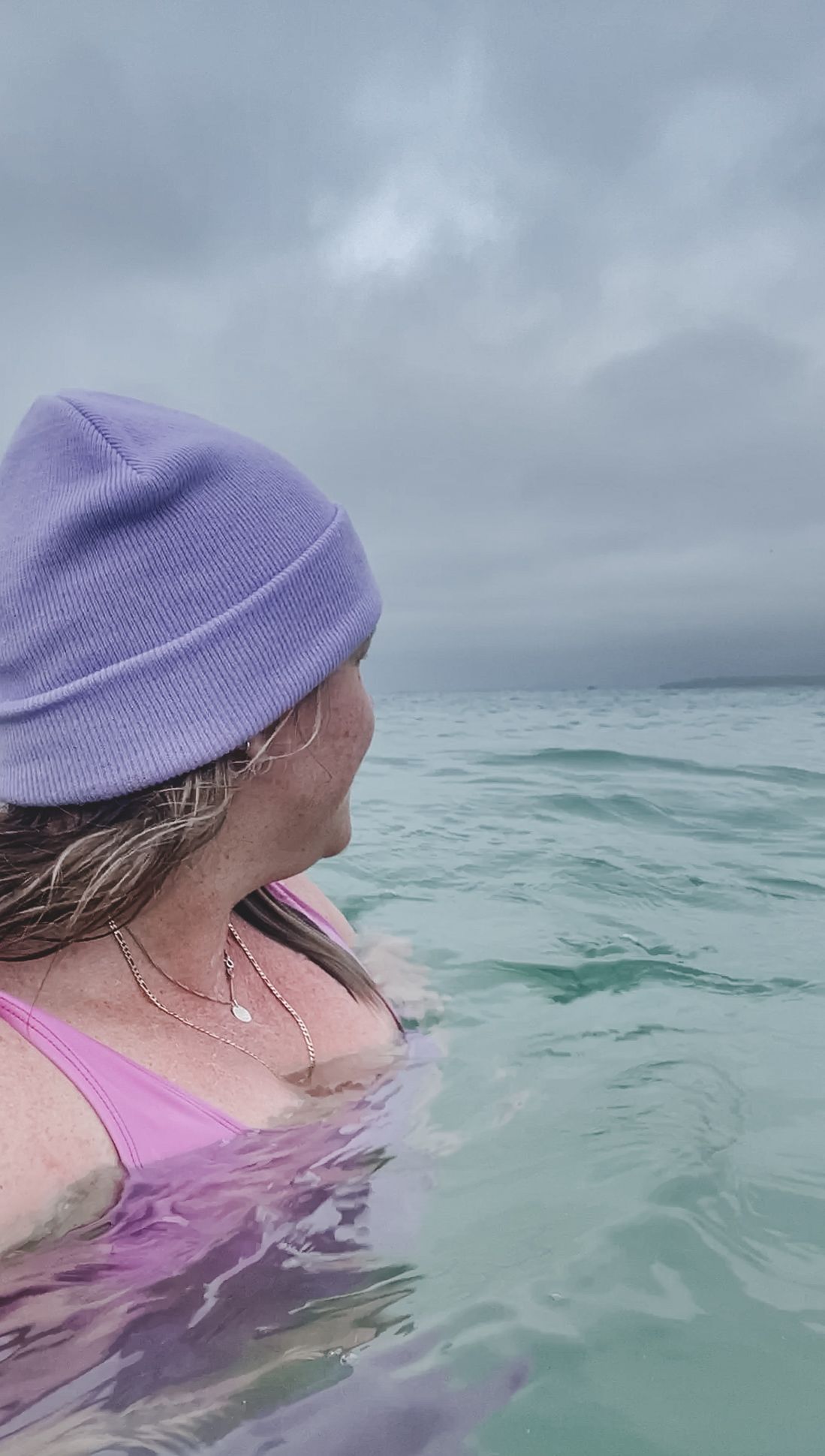 Person wearing purple beanie swimming in turquoise ocean waters on an overcast day.