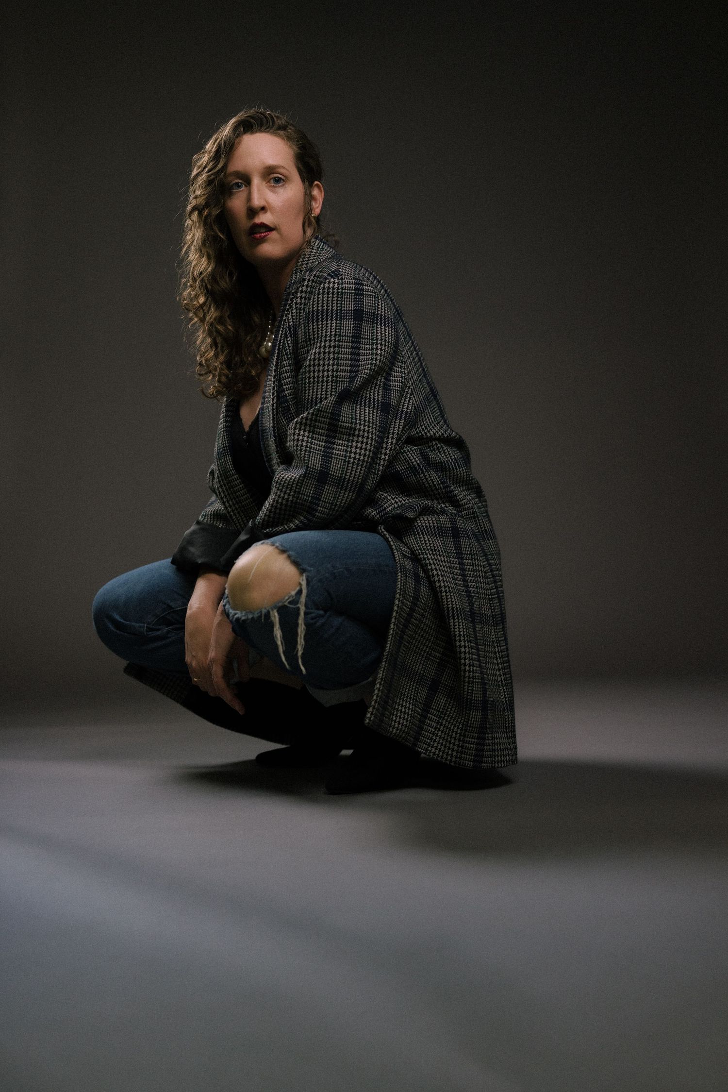 Moody studio portrait of someone wearing a plaid coat and jeans in a crouching pose.