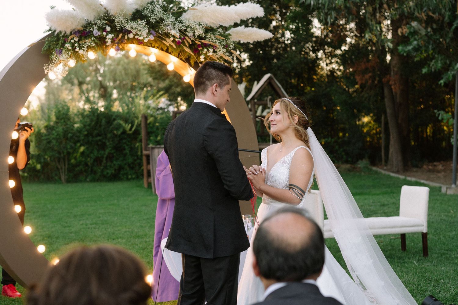 A couple shares their wedding vows during an intimate outdoor ceremony under a lit circular arch.