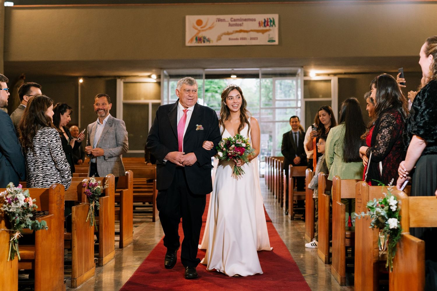 A wedding processional in a church sanctuary with floral decorations along the pews.