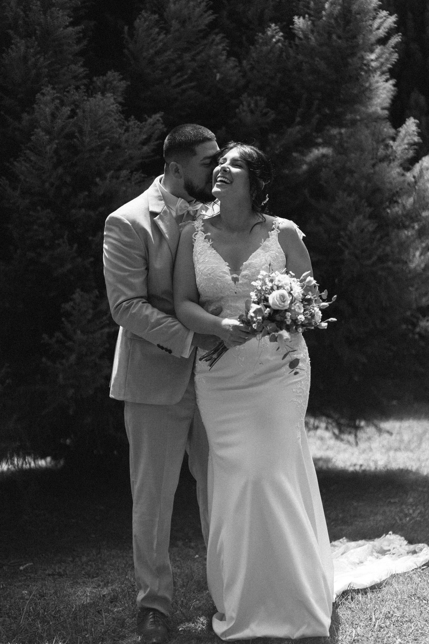 Black and white photo of a smiling bride holding bouquet and groom embracing outdoors near evergreen trees.