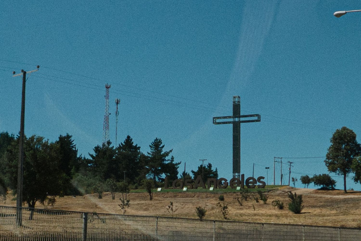 A large wooden cross stands on a hilltop against a bright blue sky with power lines in view.