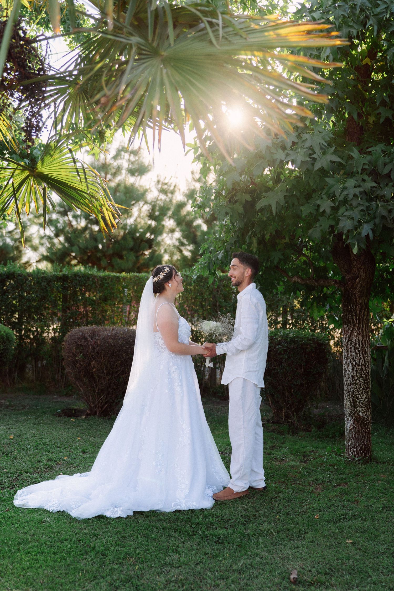 A bride and groom hold hands in a sunlit garden, surrounded by lush tropical trees on their wedding day.