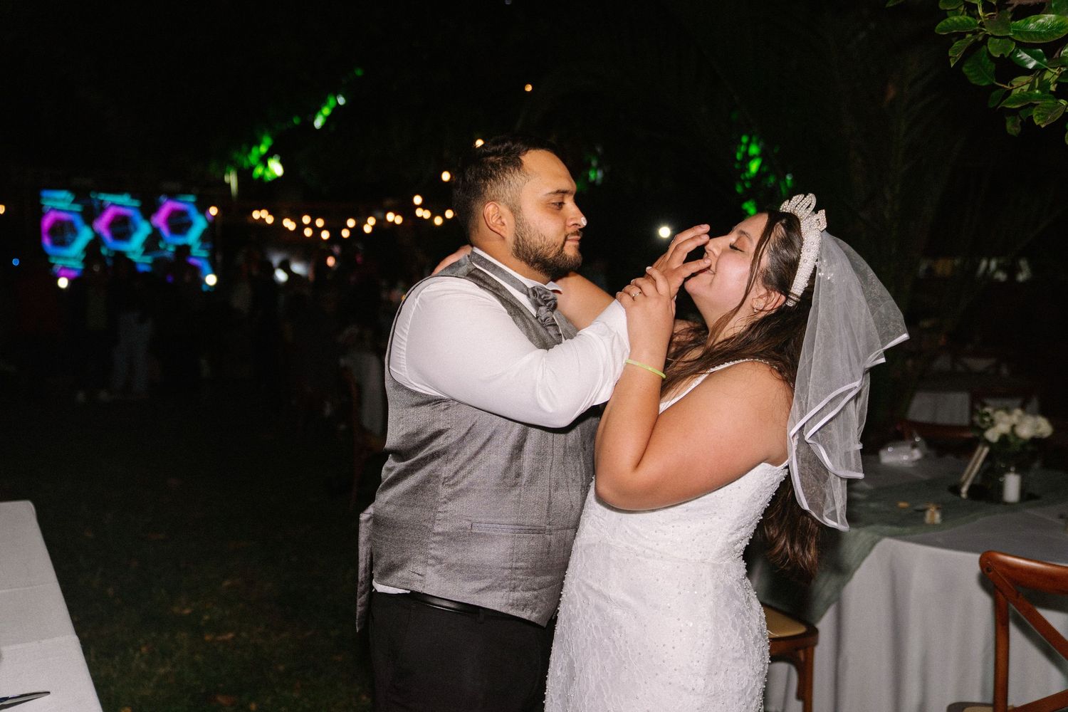 Bride and groom share cake cutting moment at outdoor evening wedding reception with festive lights.
