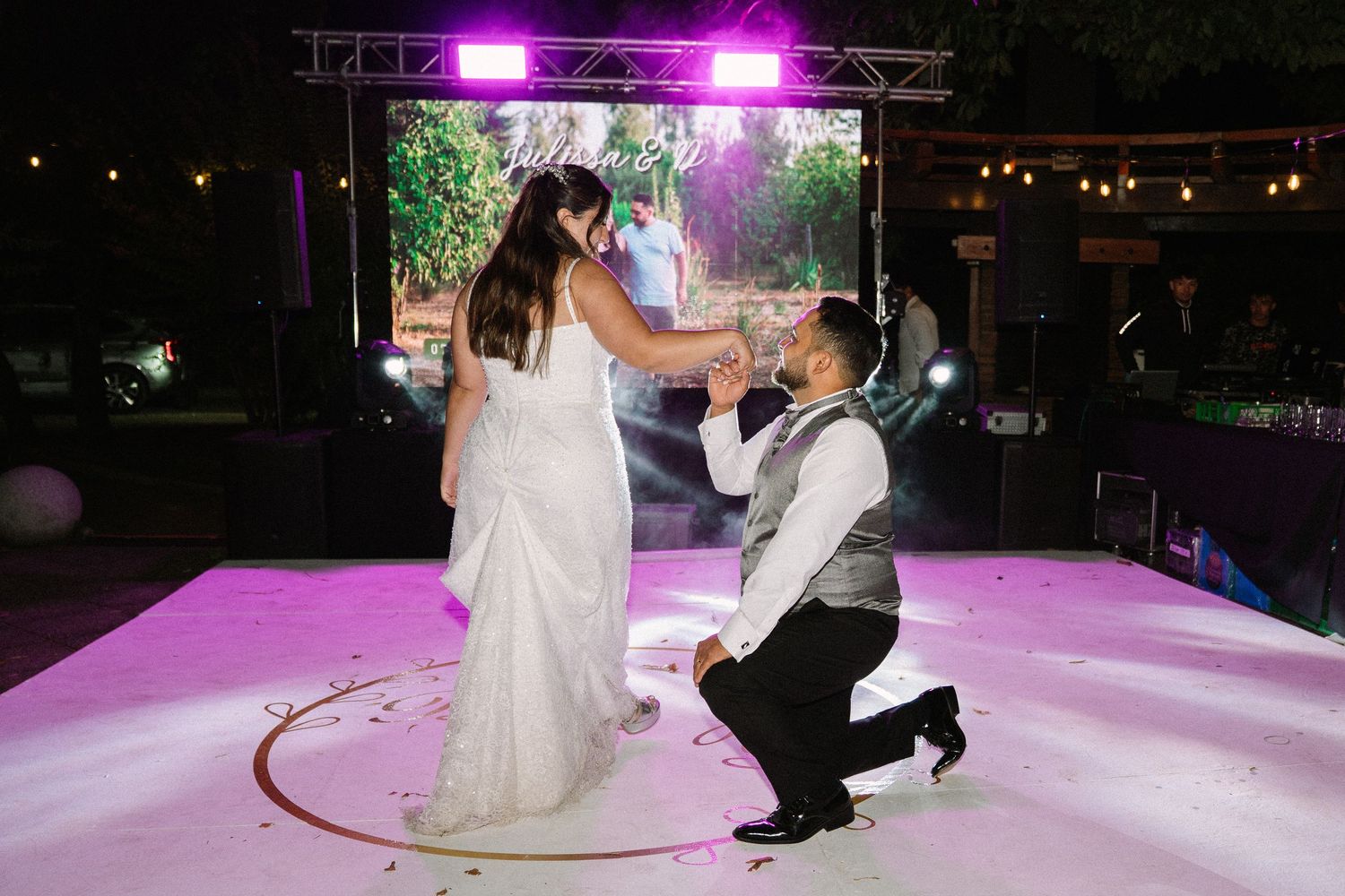 Groom kneels before bride during romantic first dance on glowing pink illuminated wedding dance floor.