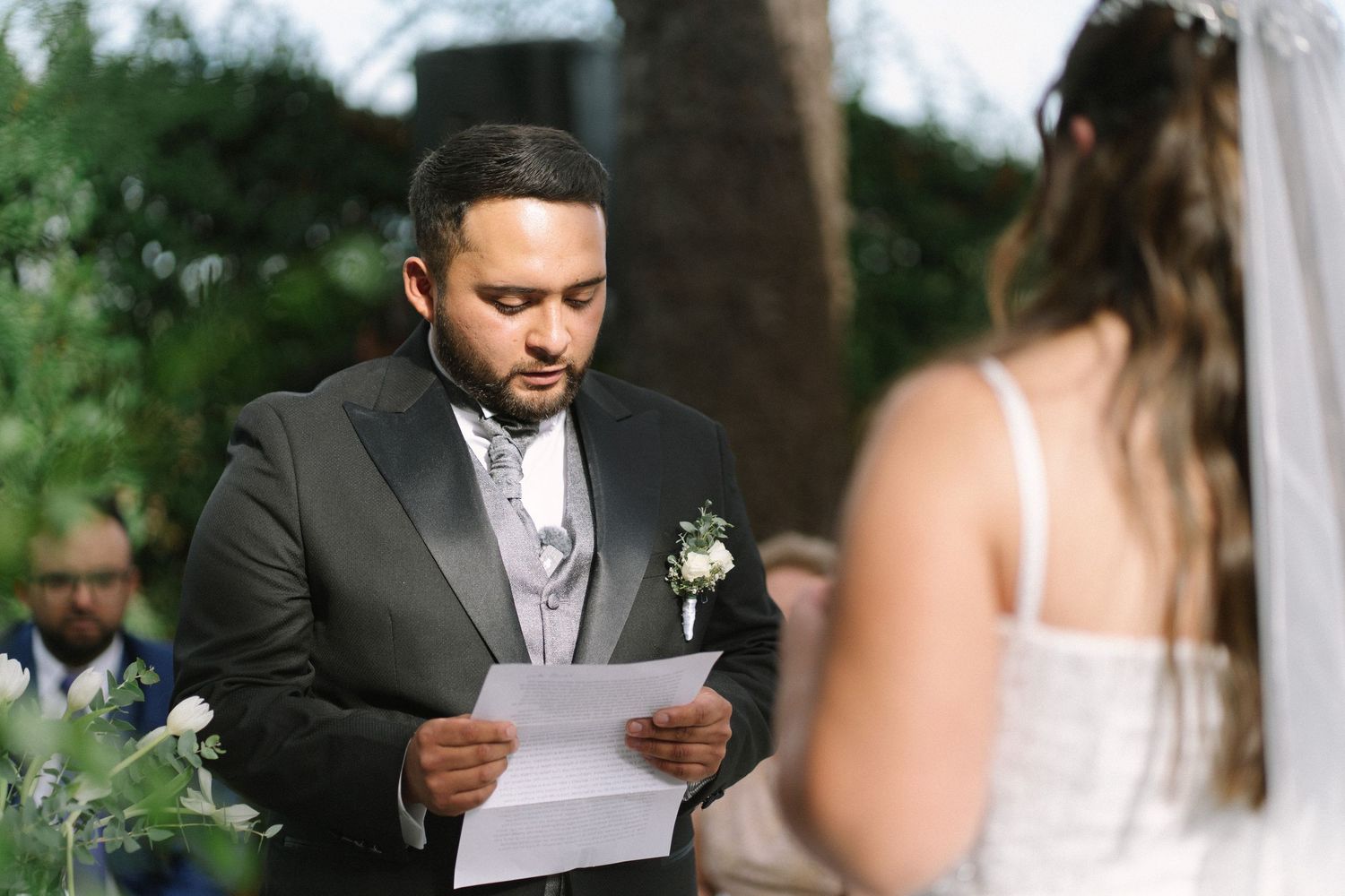 Groom in black tuxedo reading heartfelt wedding vows to bride during outdoor ceremony.