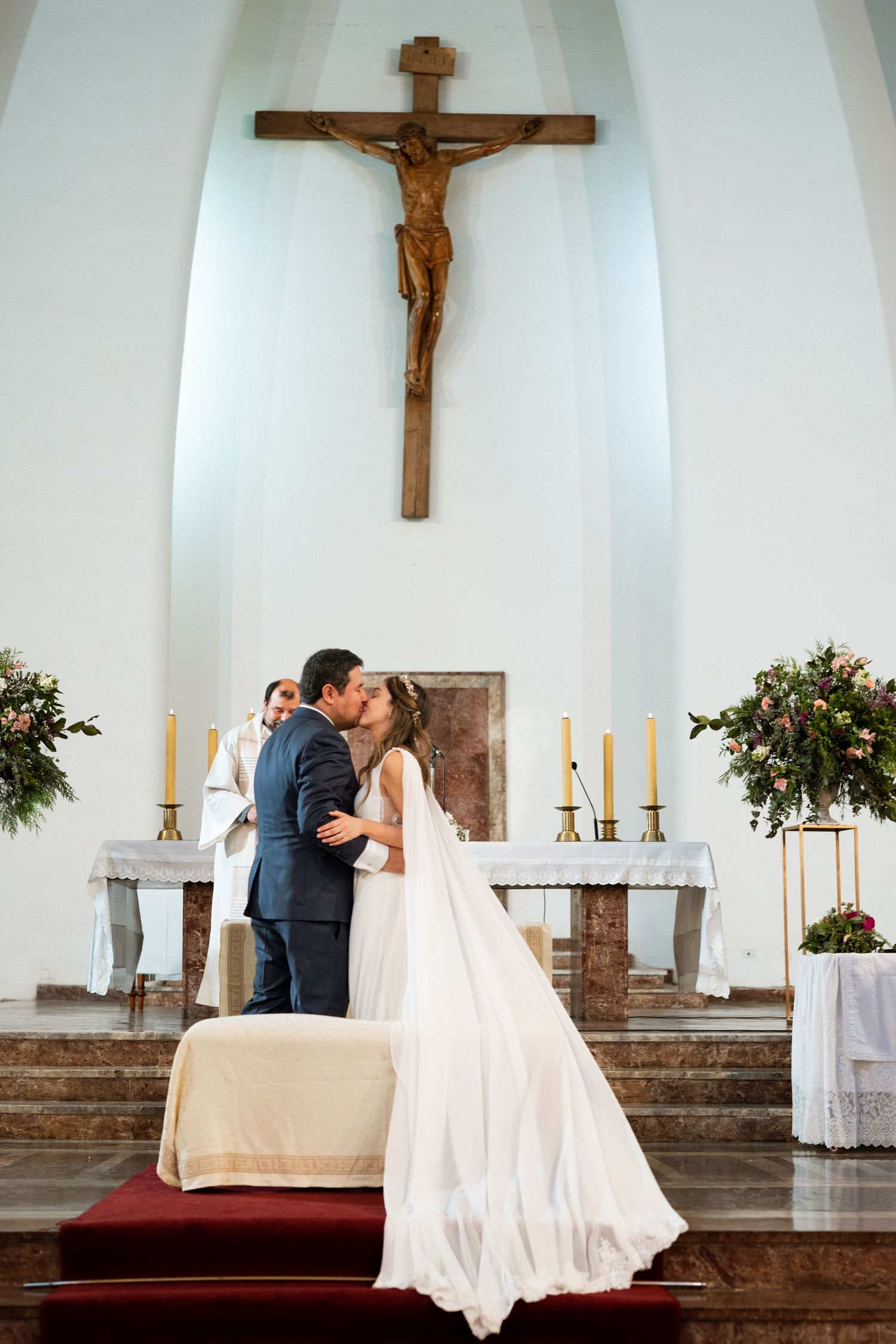 Couple shares romantic kiss during Catholic wedding ceremony with large crucifix and lit candles in church altar.