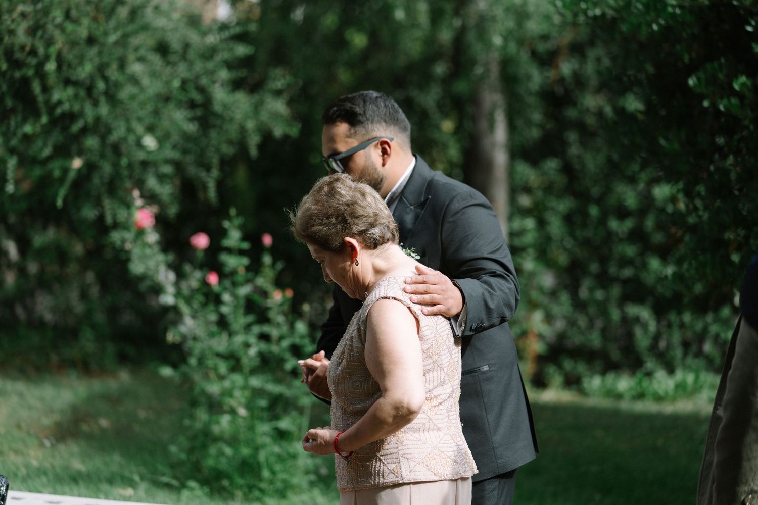A groom in a dark suit embraces an older woman in a beige dress during an outdoor wedding ceremony surrounded by lush greenery.