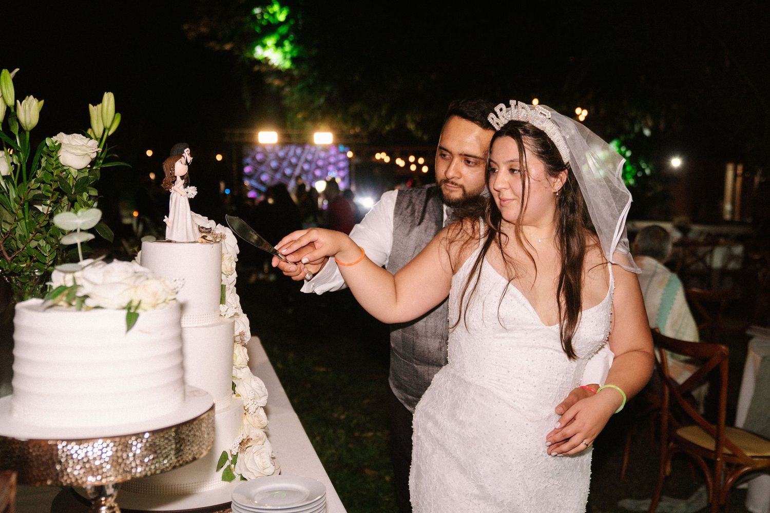 Bride and groom cutting their white tiered wedding cake together at an outdoor night reception.