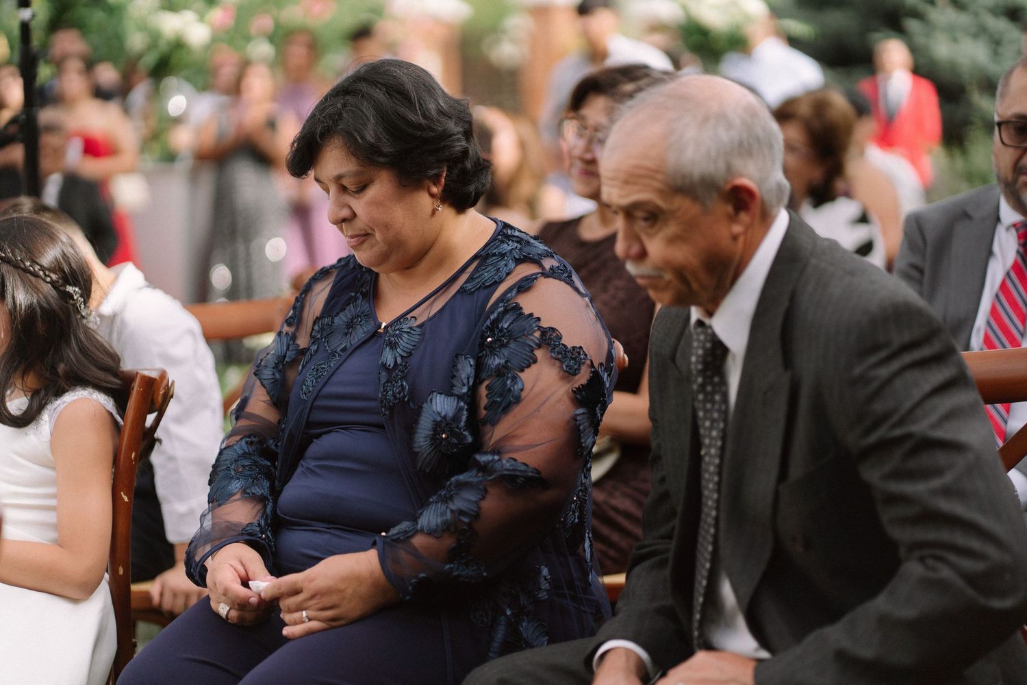 Elderly couple seated as wedding guests, woman in navy lace dress and man in suit at outdoor ceremony.