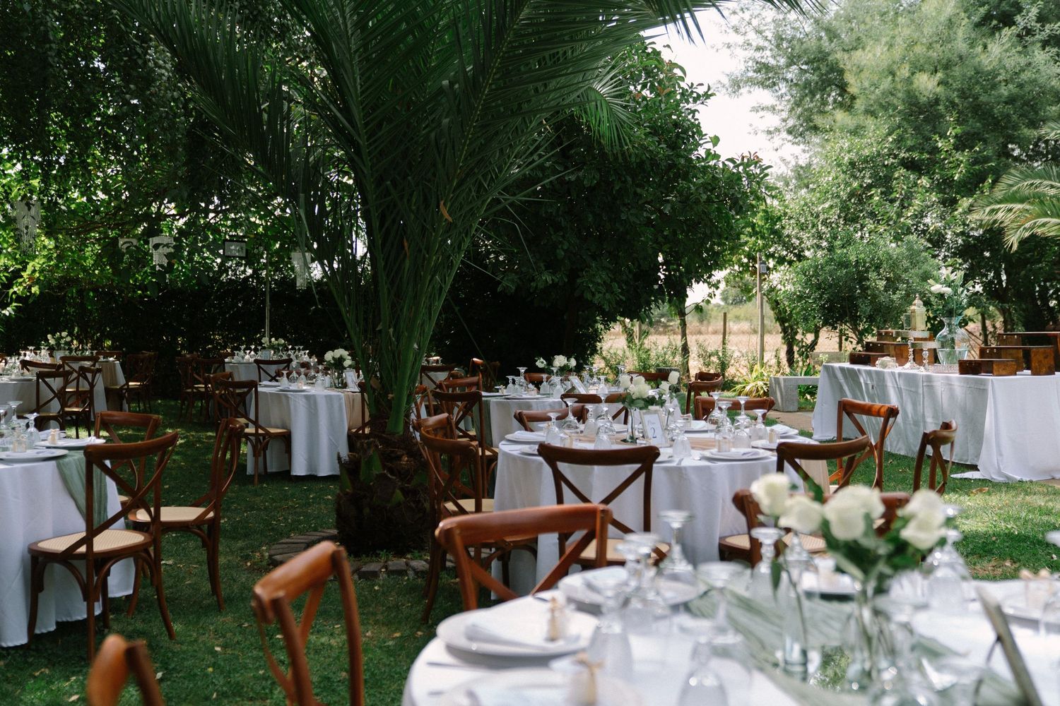 Outdoor garden wedding reception with round tables, white linens, wooden cross-back chairs, and lush tropical greenery.