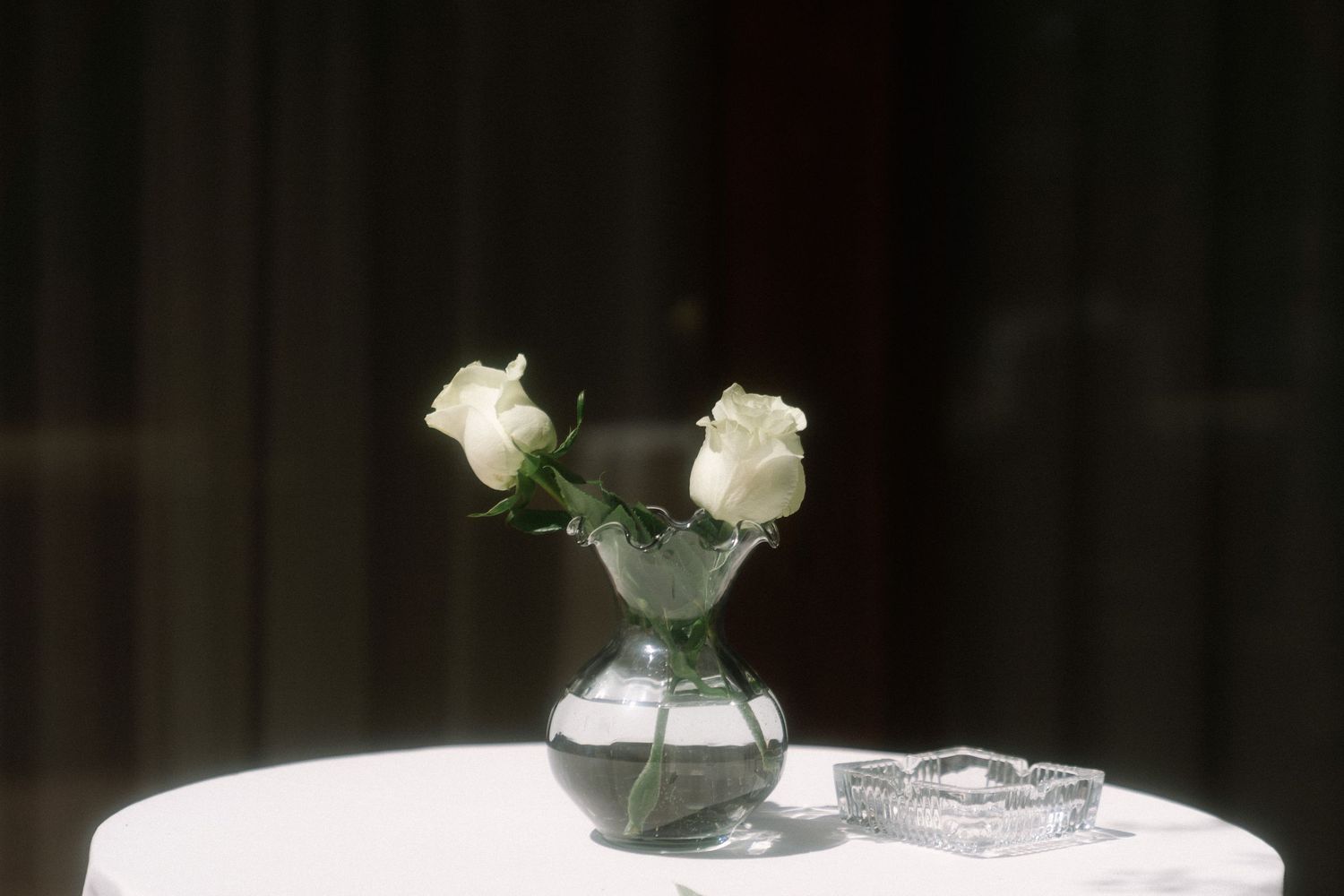 Two white roses in a small glass vase on a white tablecloth, with a crystal ashtray beside them.