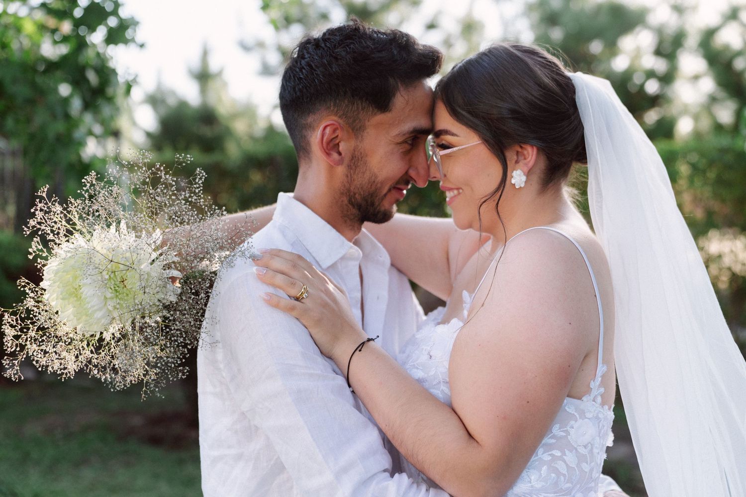 Bride and groom touch foreheads romantically holding white bouquet at outdoor wedding.