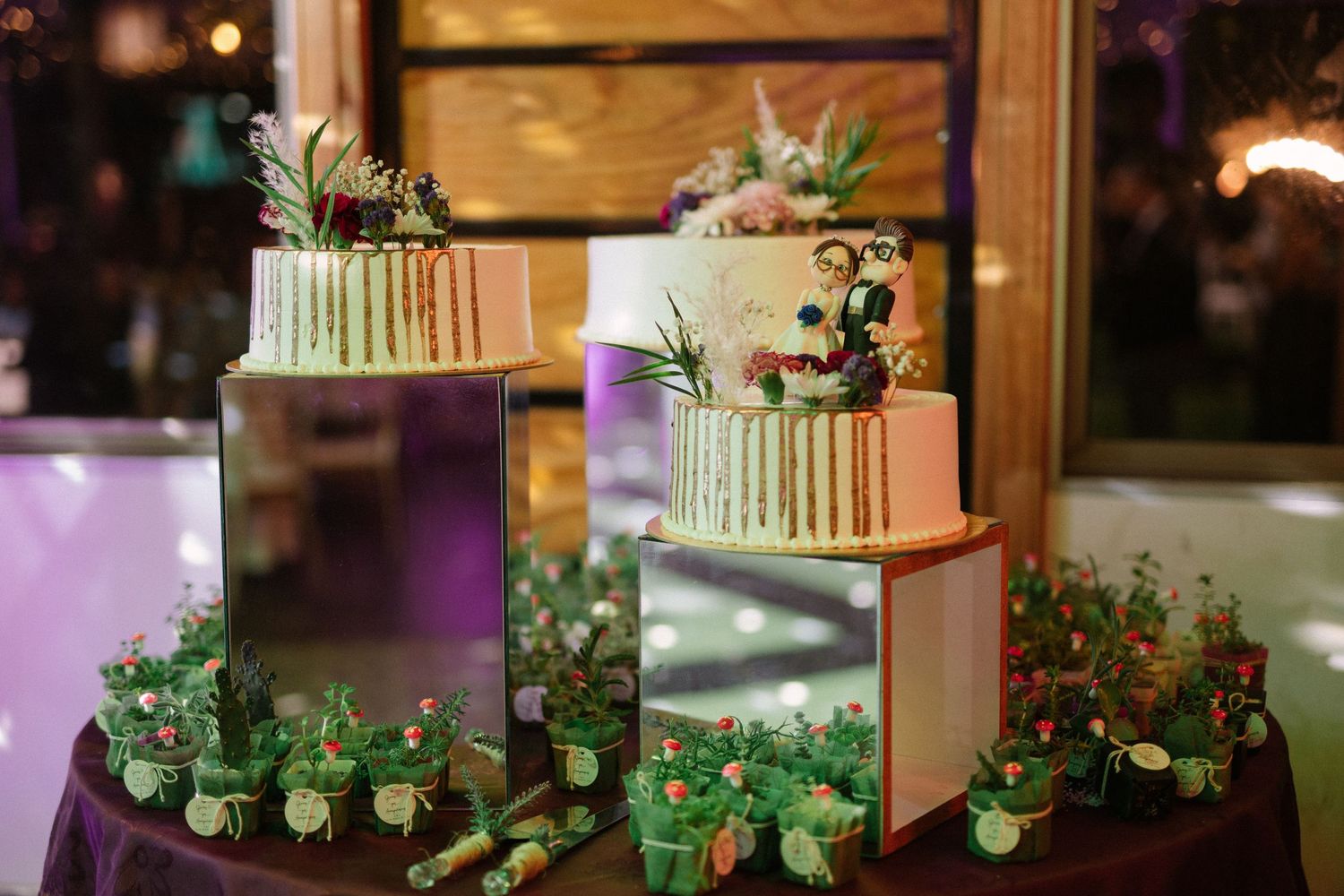 Modern tiered wedding dessert display with succulents and greenery on burgundy tablecloth.