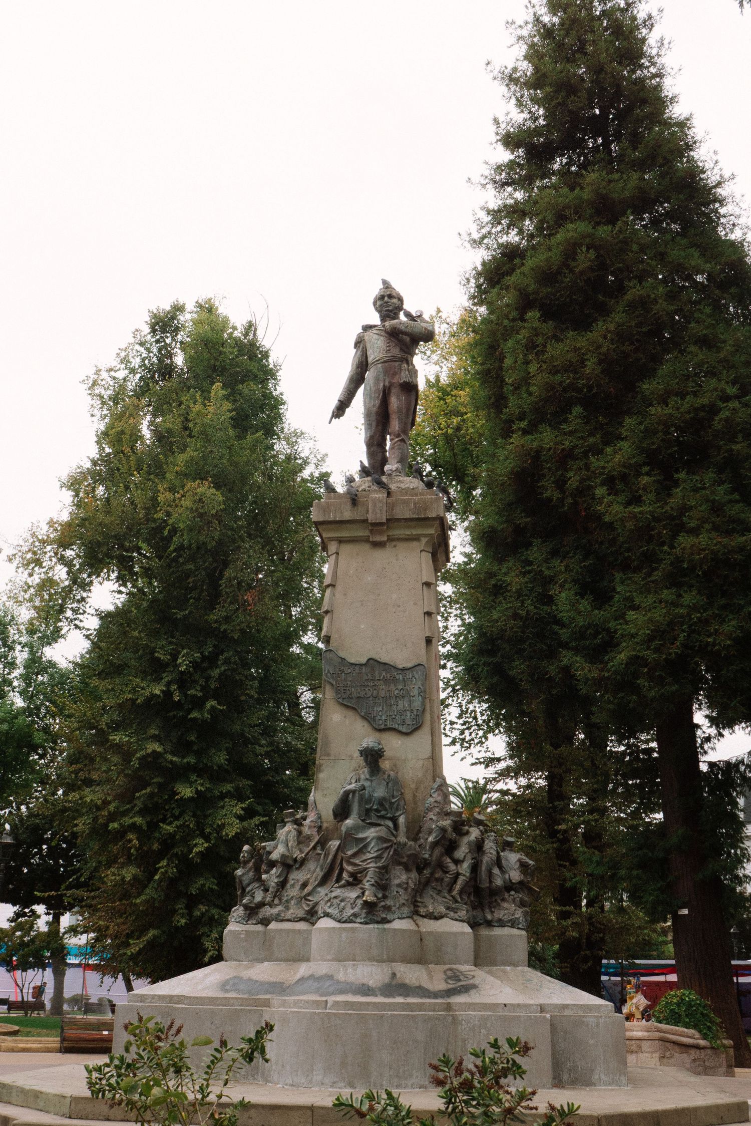 Stone monument with multiple statues and sculptural elements stands among trees in a park setting.