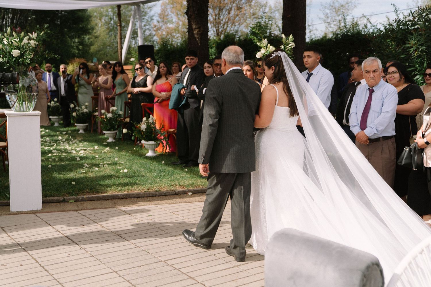 Father walking bride down outdoor wedding aisle lined with guests in garden ceremony.
