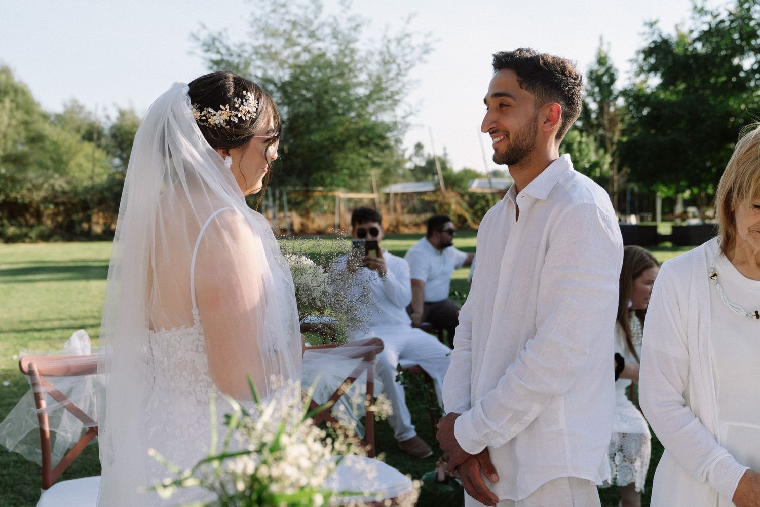 Bride in white gown and veil exchanges vows with groom in white suit during outdoor garden wedding ceremony.