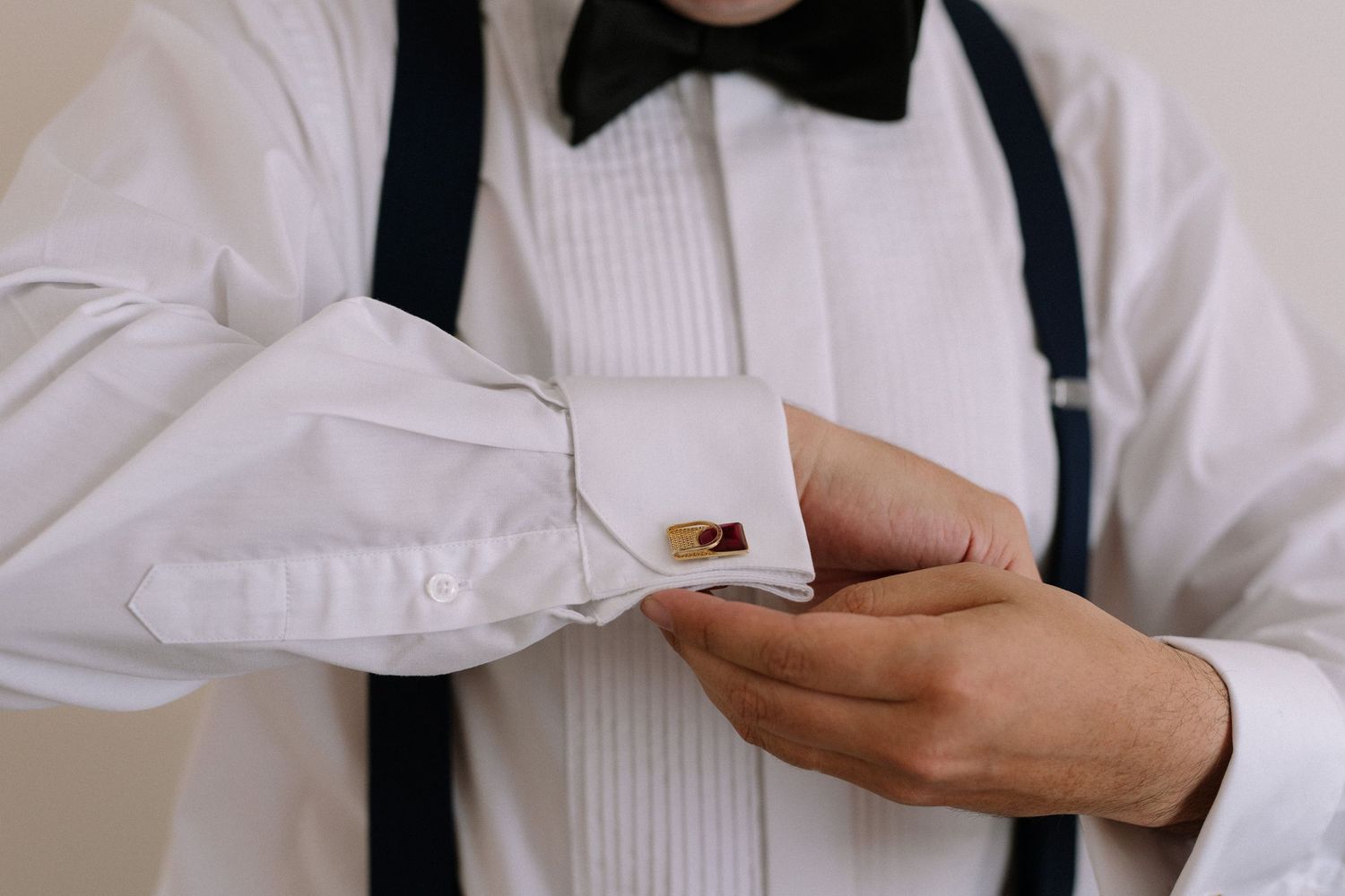 Person fastening gold cufflink on white dress shirt sleeve with suspenders visible.