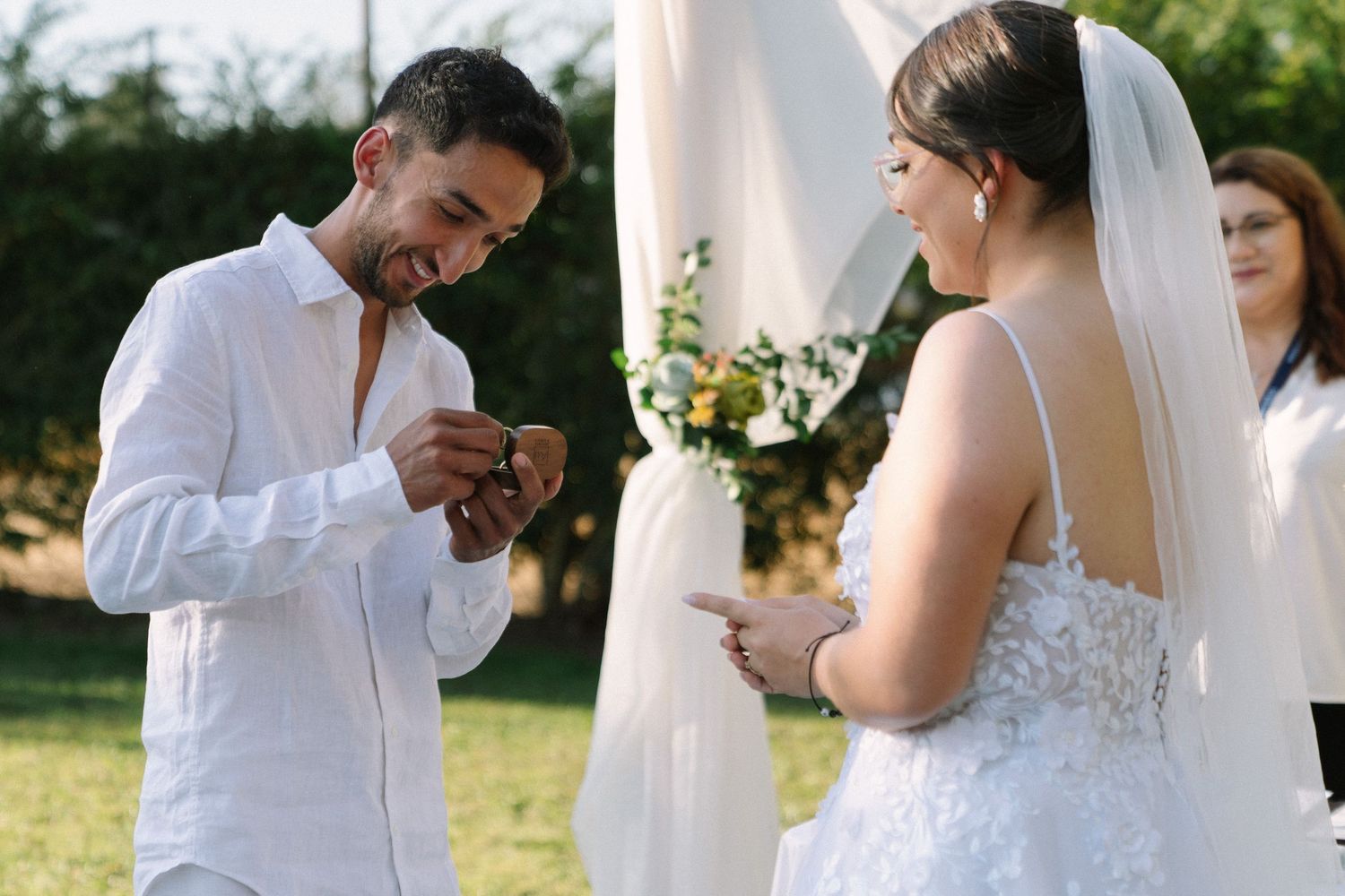 Groom placing wedding ring on bride's finger during outdoor ceremony, both dressed in white attire.