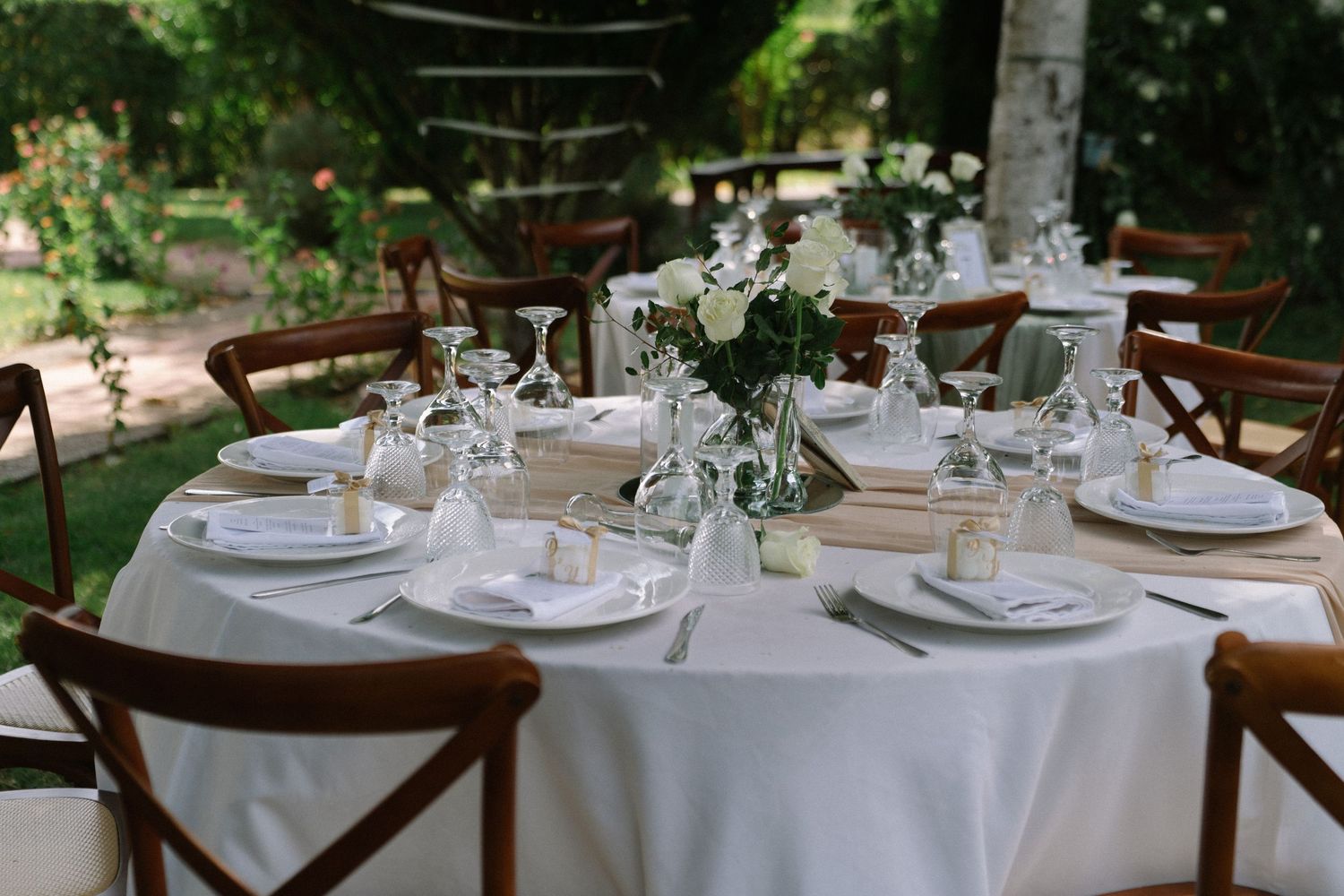 Elegantly set outdoor wedding reception table with white linens, floral centerpiece, and wooden cross-back chairs.