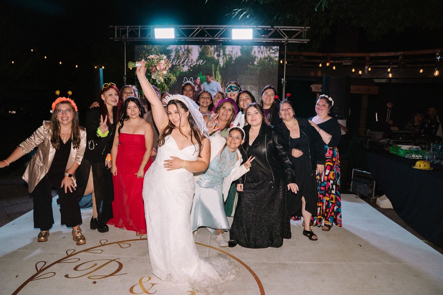 Bride raises bouquet triumphantly surrounded by group of women guests on decorated wedding dance floor.