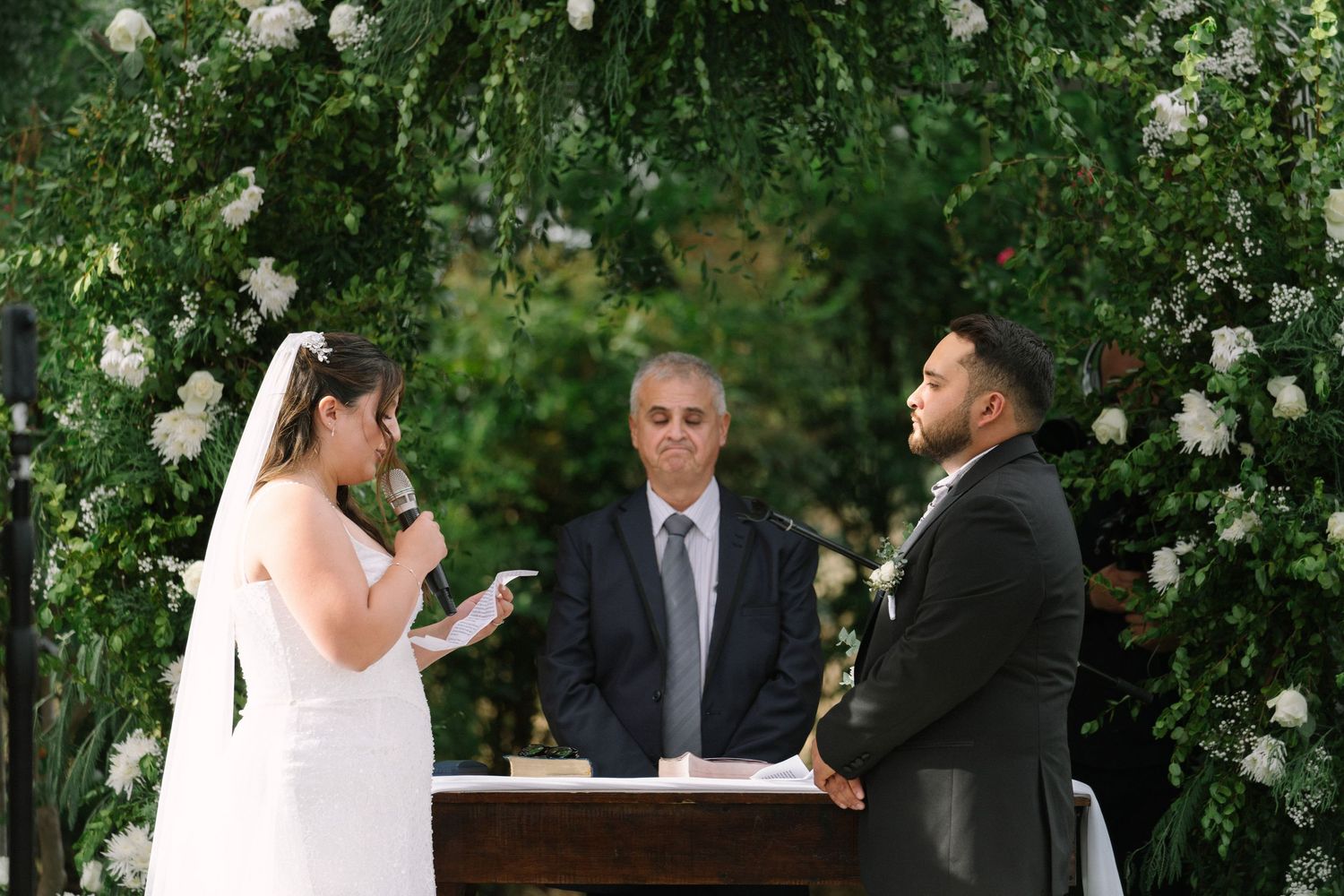 Bride reads vows to groom at outdoor wedding ceremony beneath lush green floral arch with white flowers.