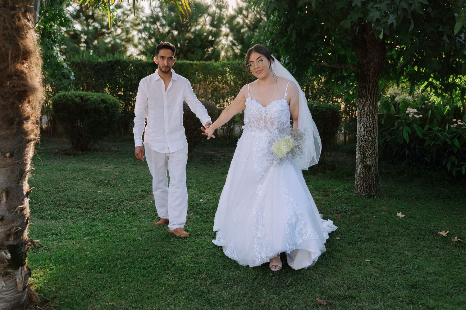 Bride in white lace gown and groom in white suit hold hands while walking through a lush garden.