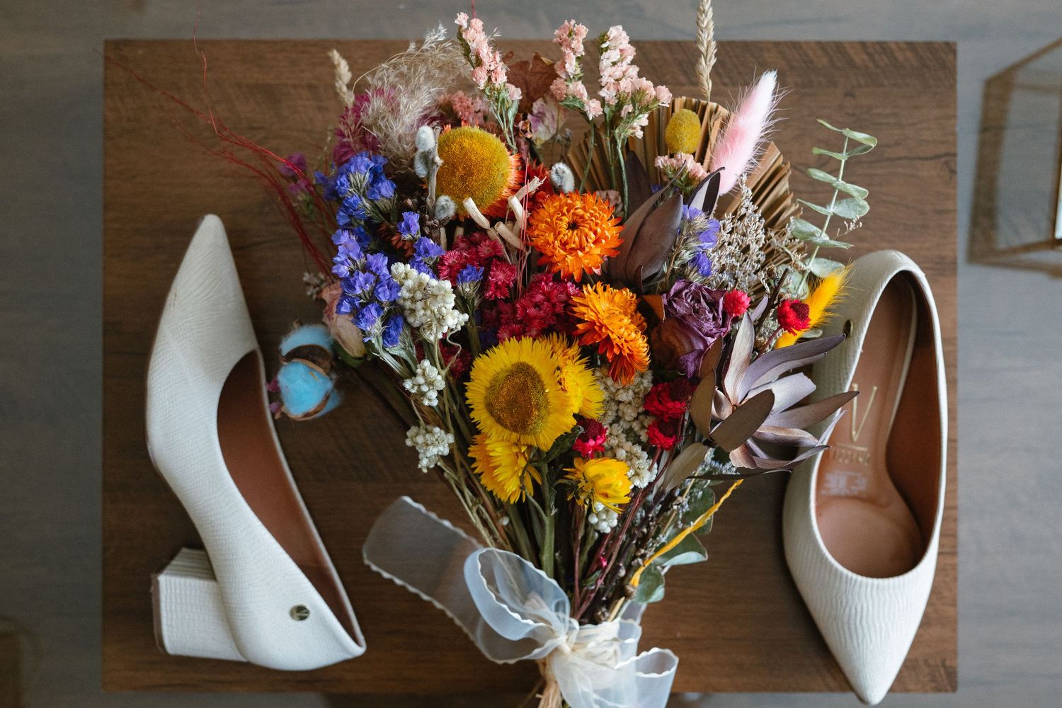 Colorful wildflower bouquet with sunflowers sits next to white bridal shoes on wooden surface.