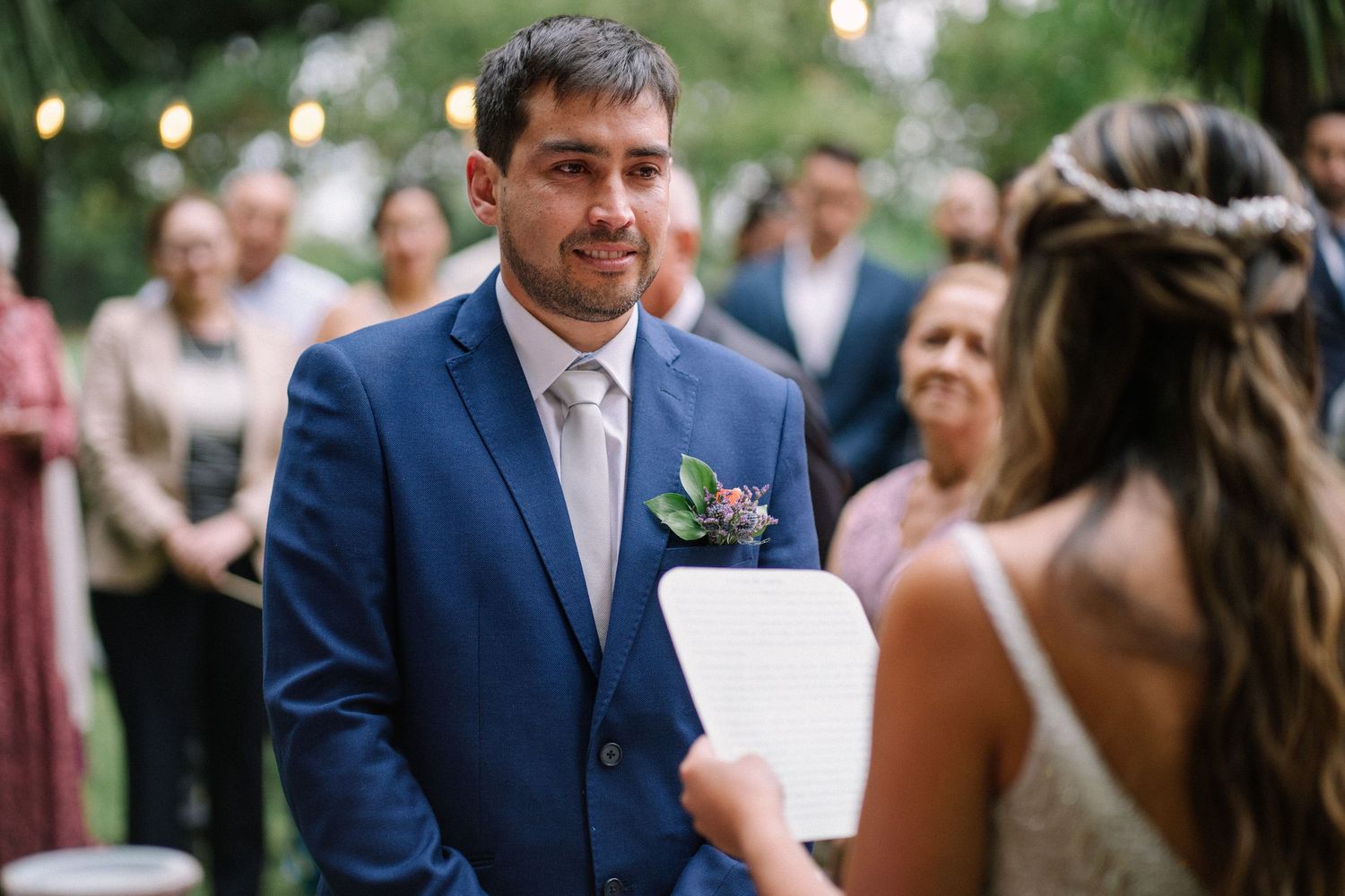 A groom in a navy blue suit smiles emotionally during an outdoor wedding ceremony.