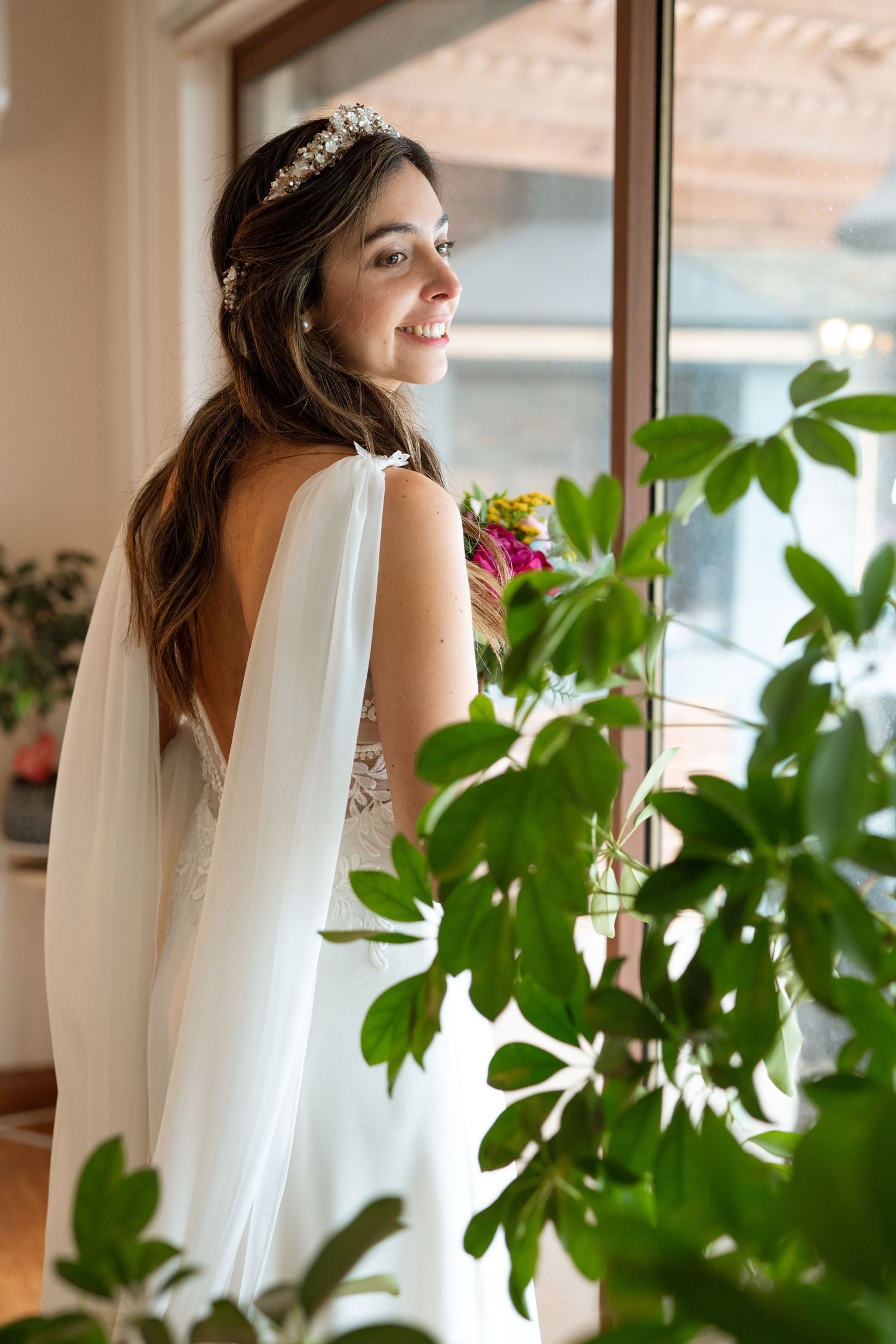 A bride in a flowing white dress with veil stands by a window surrounded by green plants.