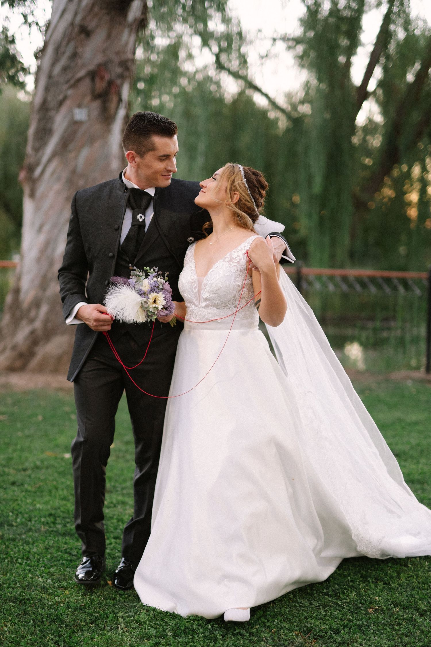 Wedding couple stands by eucalyptus trees, sharing a loving moment with purple floral bouquet.