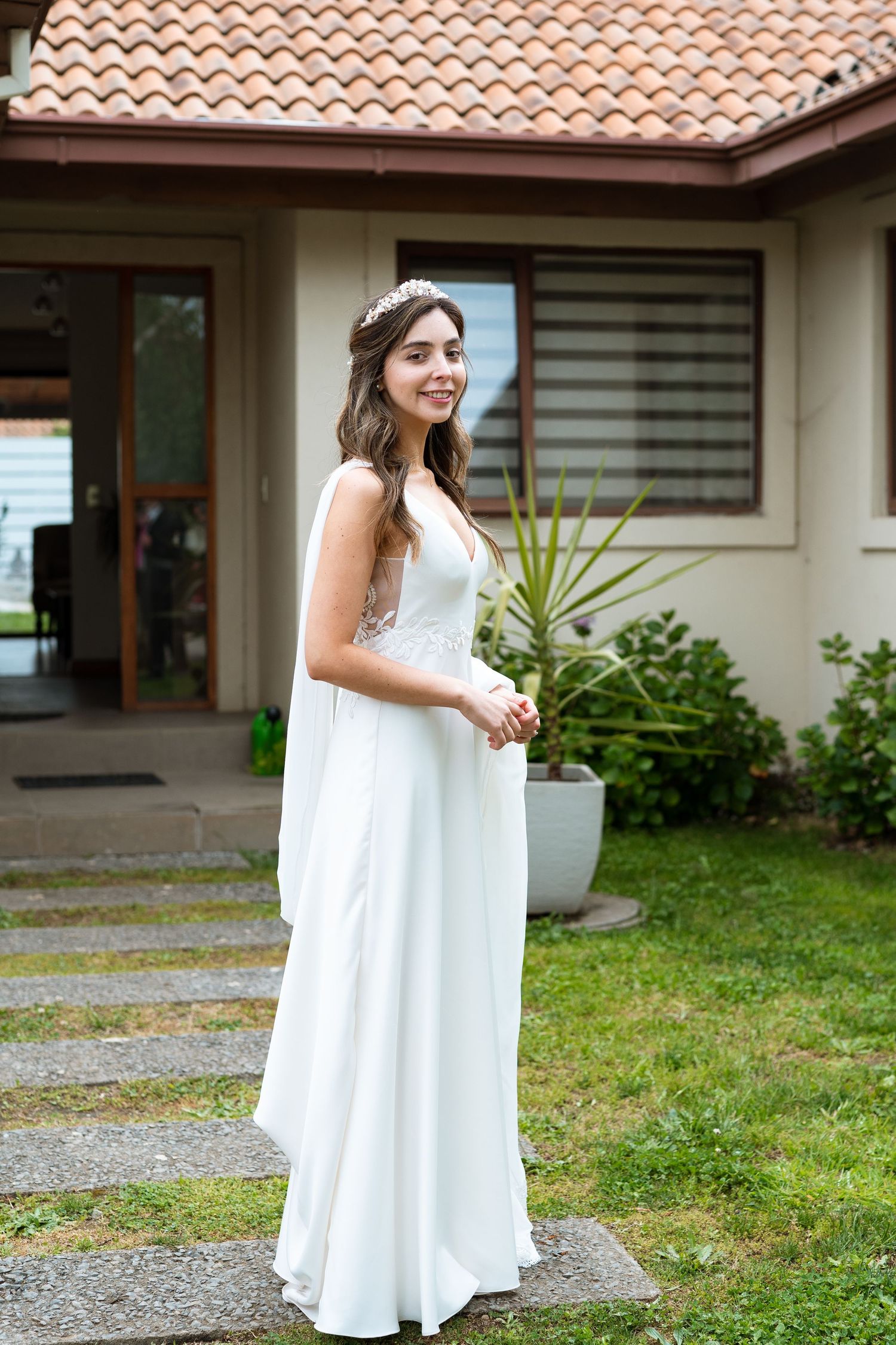 A bride in a flowing white dress stands in front of a house with terracotta roof tiles.