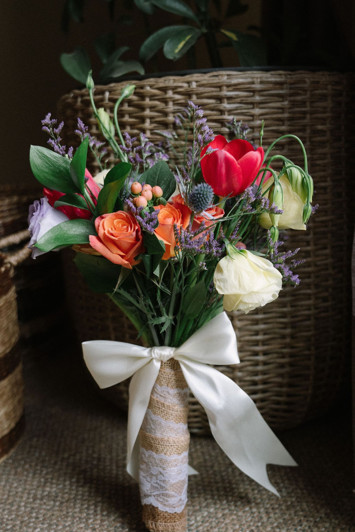 A rustic wedding bouquet with red tulips, orange roses, and white flowers tied with a white ribbon bow and burlap wrap.