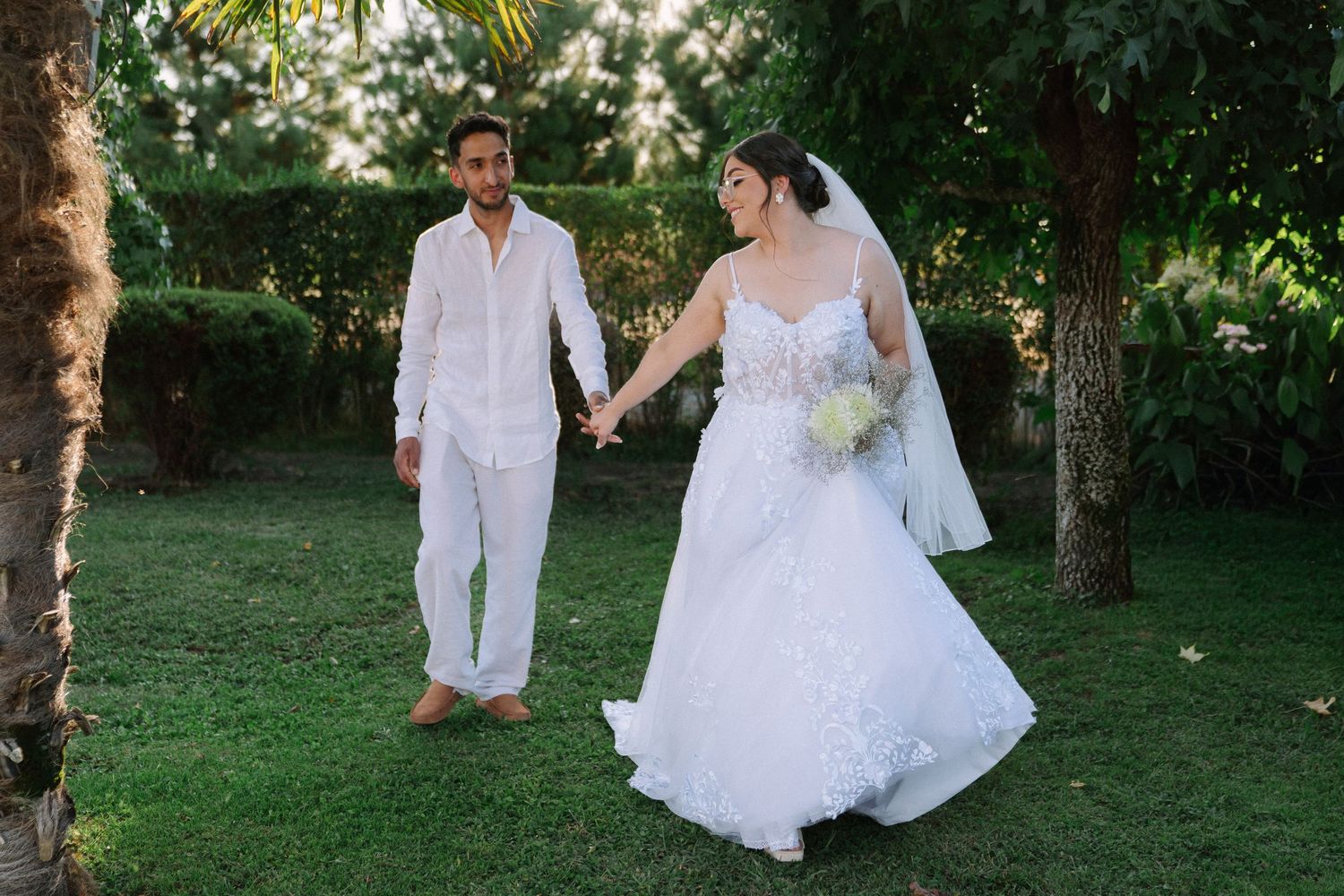 Newlyweds stroll hand in hand across green lawn surrounded by tropical trees.