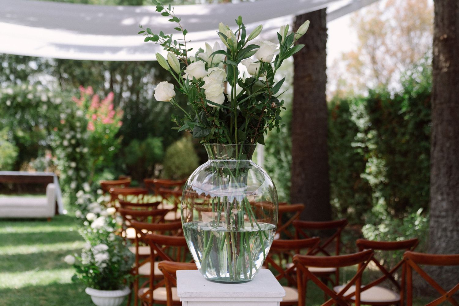 Glass vase with white roses and greenery on white pedestal at an outdoor wedding ceremony with wooden chairs.