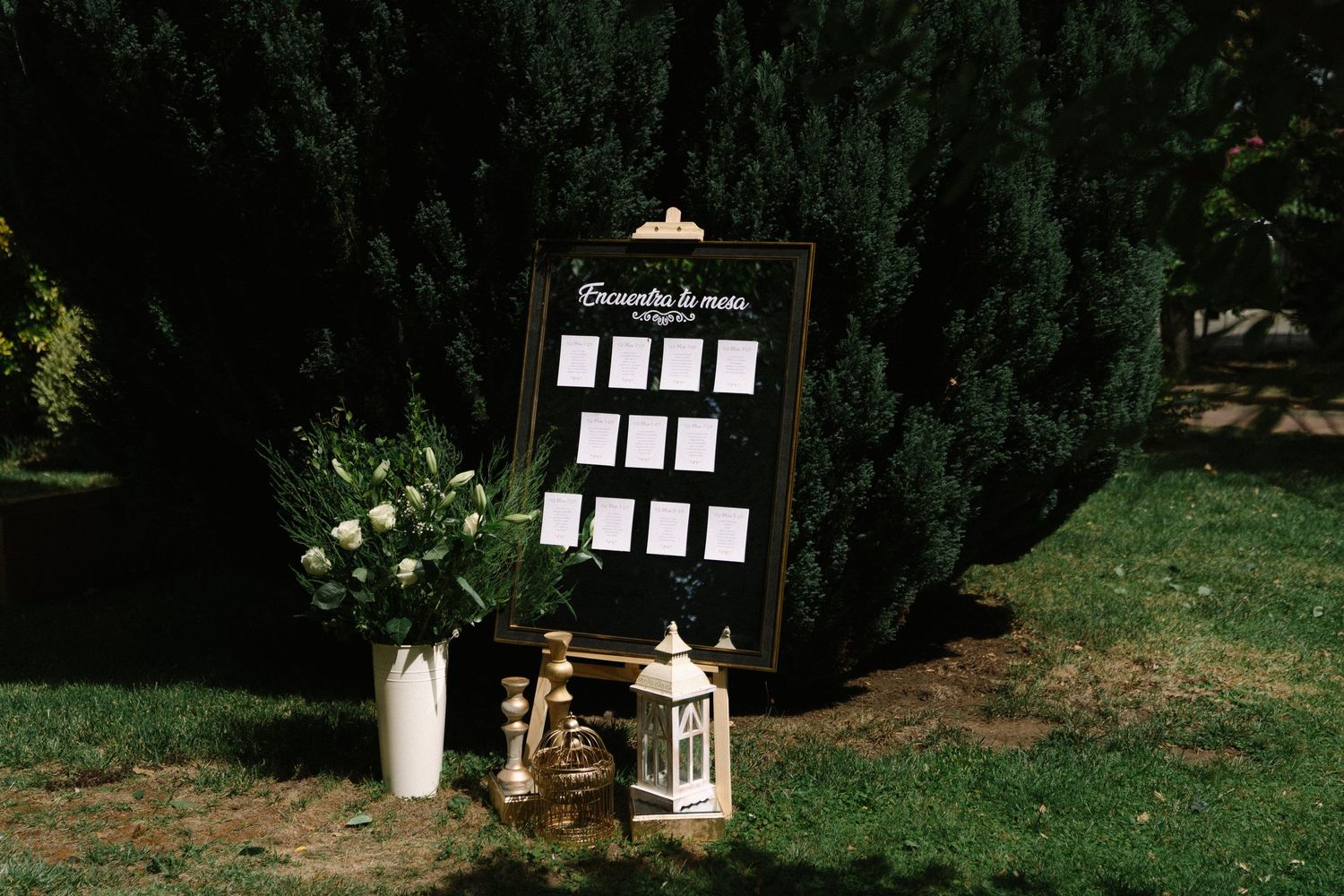 Elegant wedding seating chart displayed on a black framed board surrounded by white flowers and lanterns.