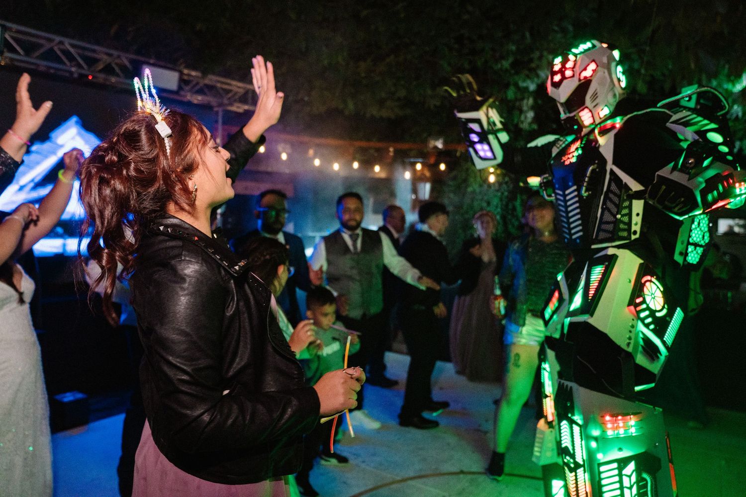 Woman in leather jacket dances with illuminated robot entertainer at outdoor night party event.