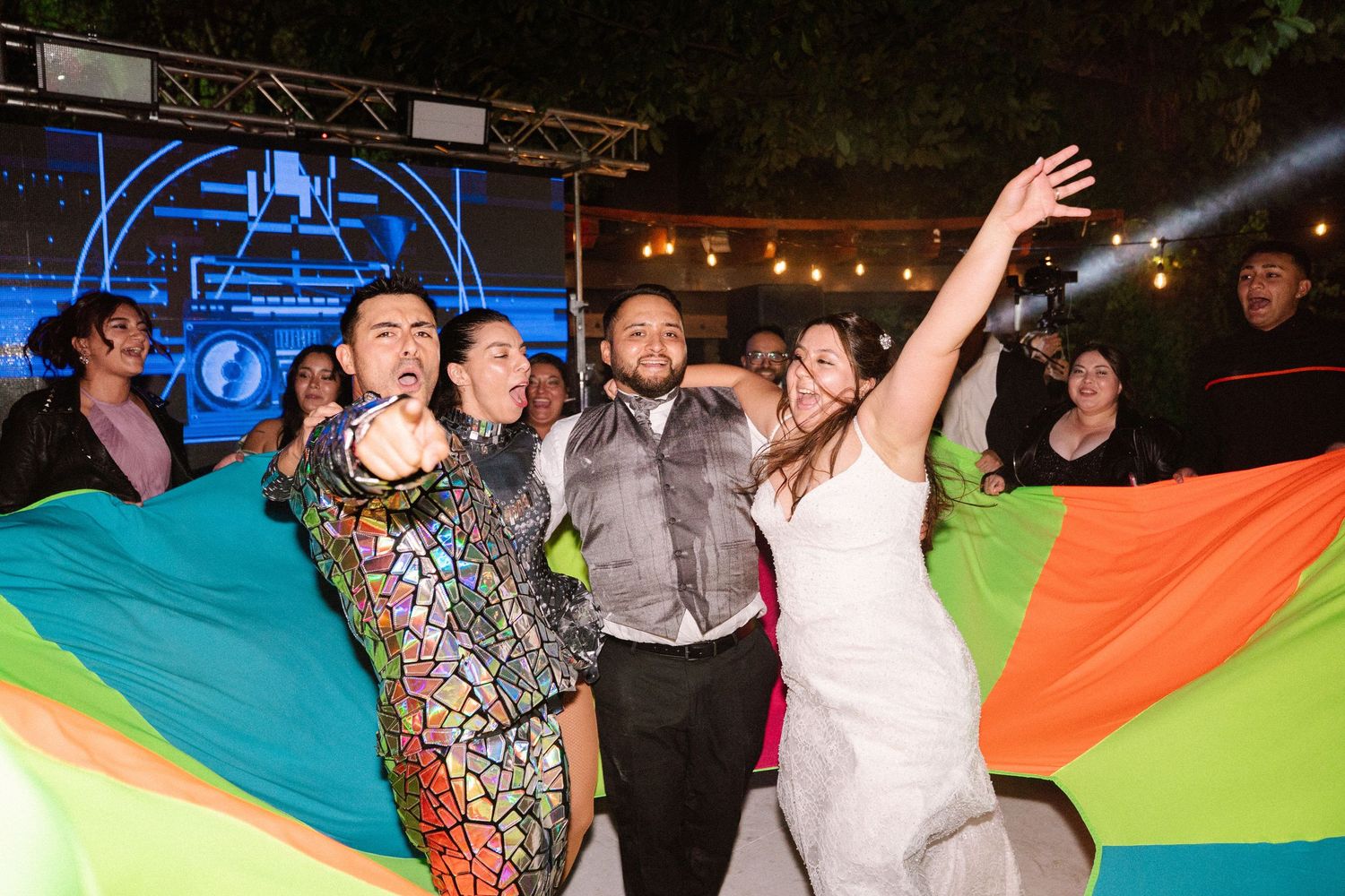 Bride, groom and guests celebrate joyfully with colorful rainbow flag at vibrant outdoor wedding reception.
