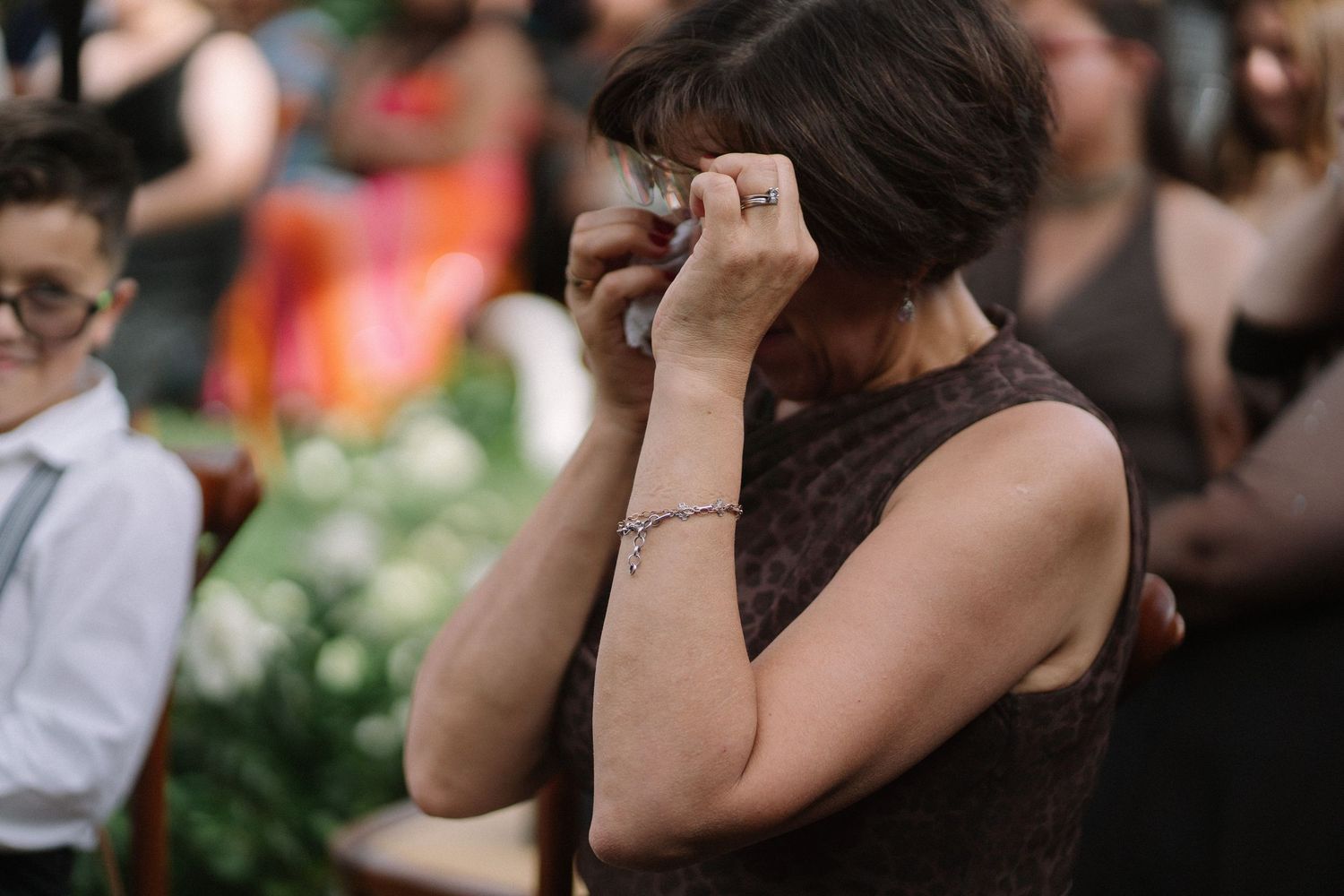A woman in a brown lace dress wipes tears from her eyes during an emotional wedding ceremony.