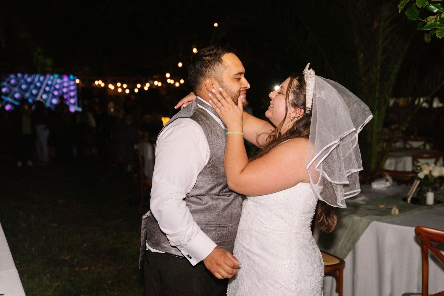 Bride and groom share romantic kiss under string lights during intimate outdoor nighttime wedding reception.