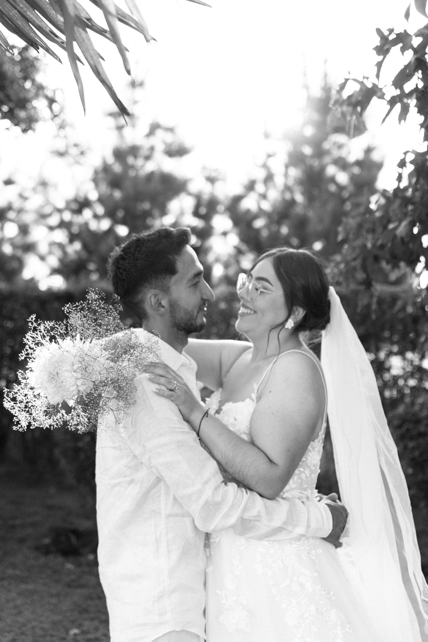 Black and white photo of a smiling bride and groom embracing outdoors on their wedding day.