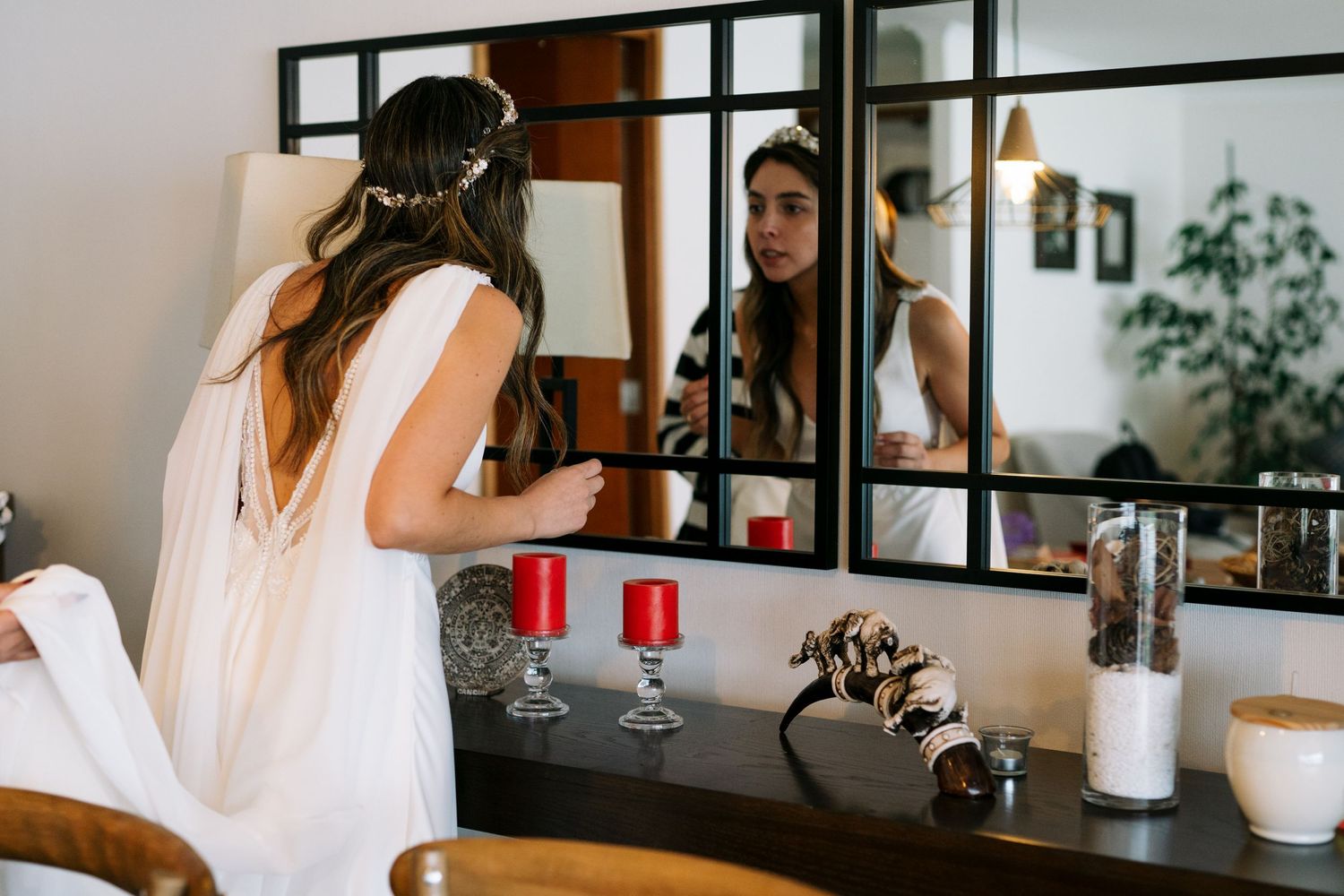 A back view of a bride in white dress looking at her reflection in a vanity mirror with candles.