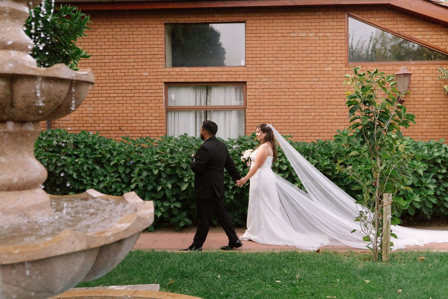 Bride and groom walk hand in hand on green lawn beside brick building and stone fountain.