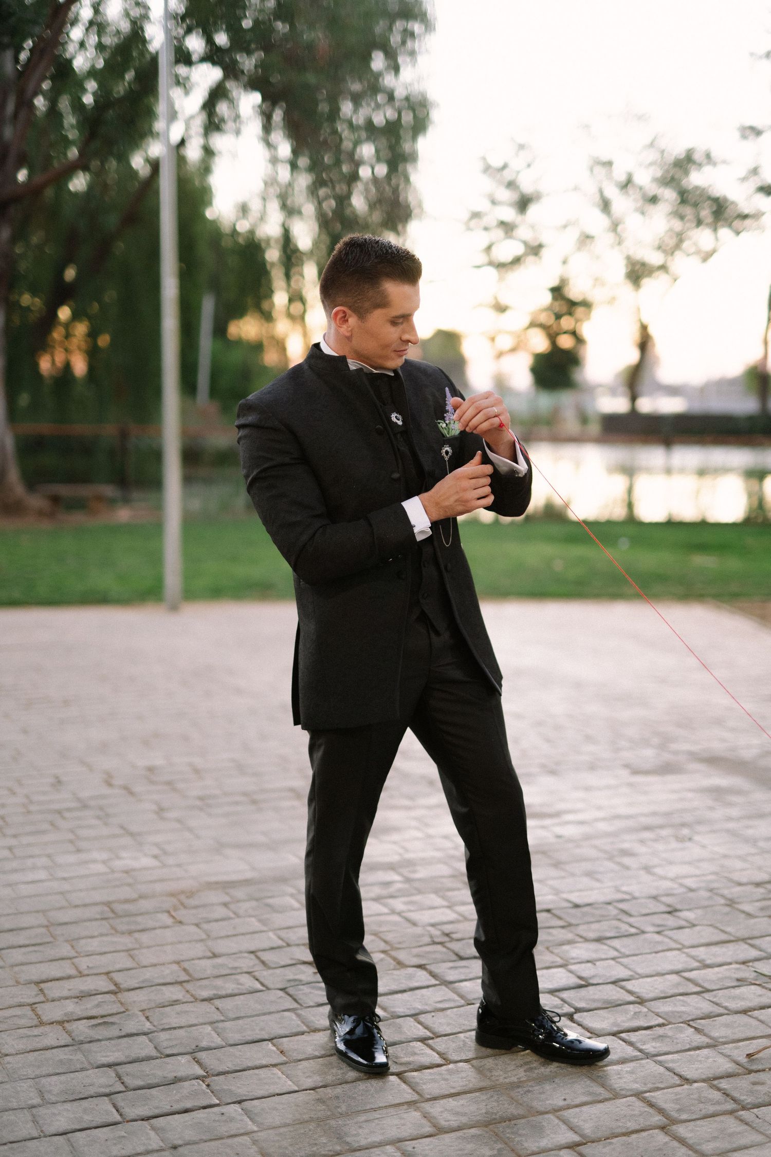 A groom in classic black tuxedo adjusts his cufflinks on a stone pathway.