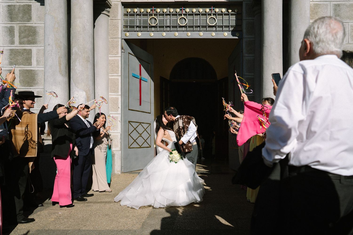 Wedding party and guests celebrate outside church entrance with bubbles and confetti.
