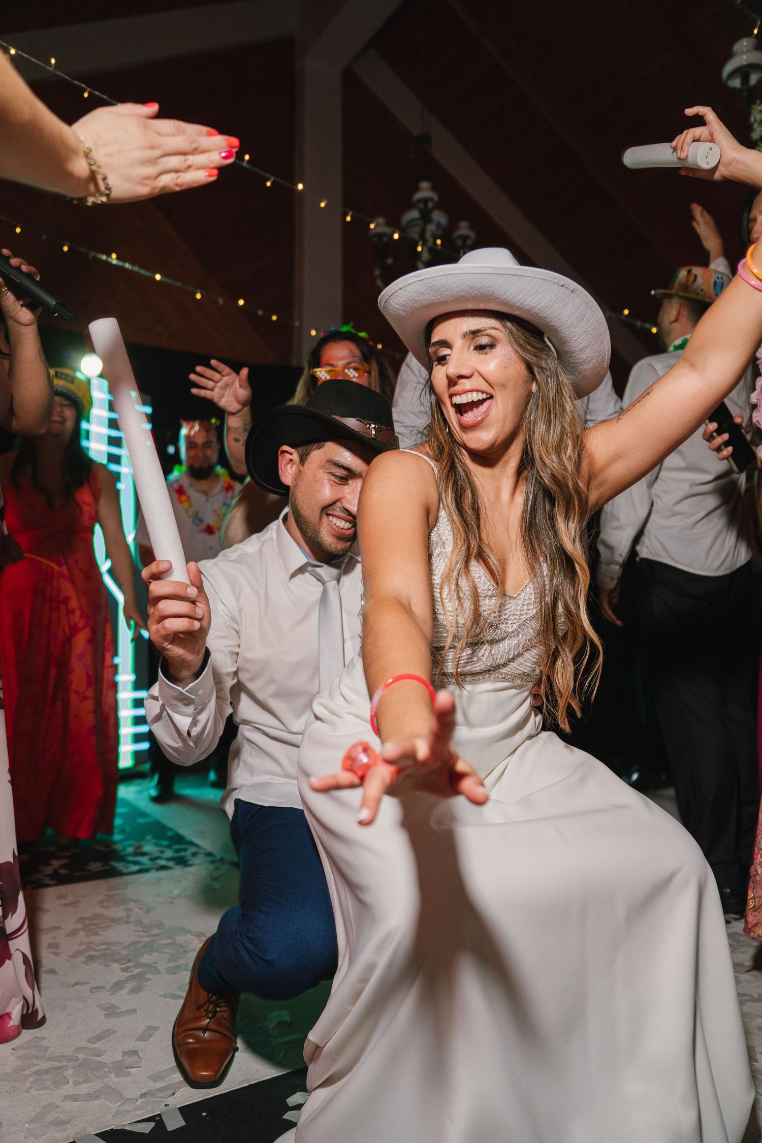 Wedding reception dance party with guests wearing cowboy hats and dancing under string lights.
