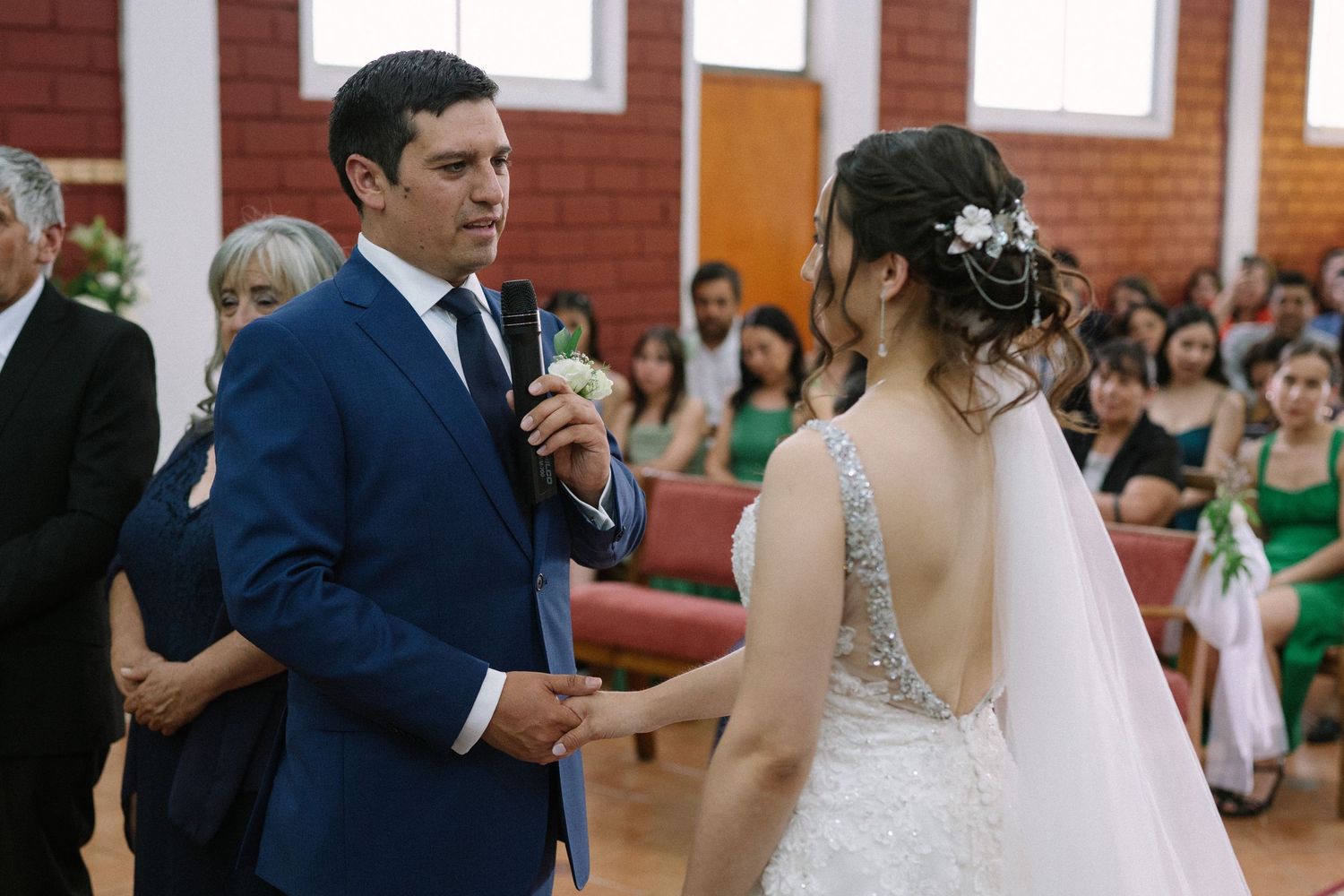 Bride and groom exchange rings during indoor wedding ceremony with guests seated in background.